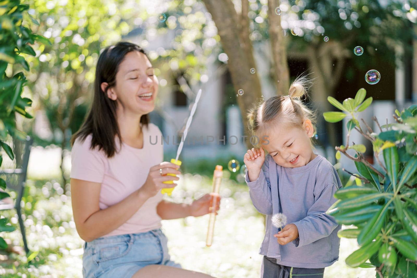 Laughing mother blowing bubbles at squinting smiling little girl in green garden. High quality photo
