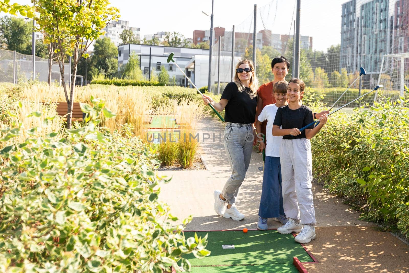 A woman is teaching her daughter to play golf. They are standing on the golf course, the girl is holding a golf club, the woman is standing behind her and directs her to strike. High quality photo