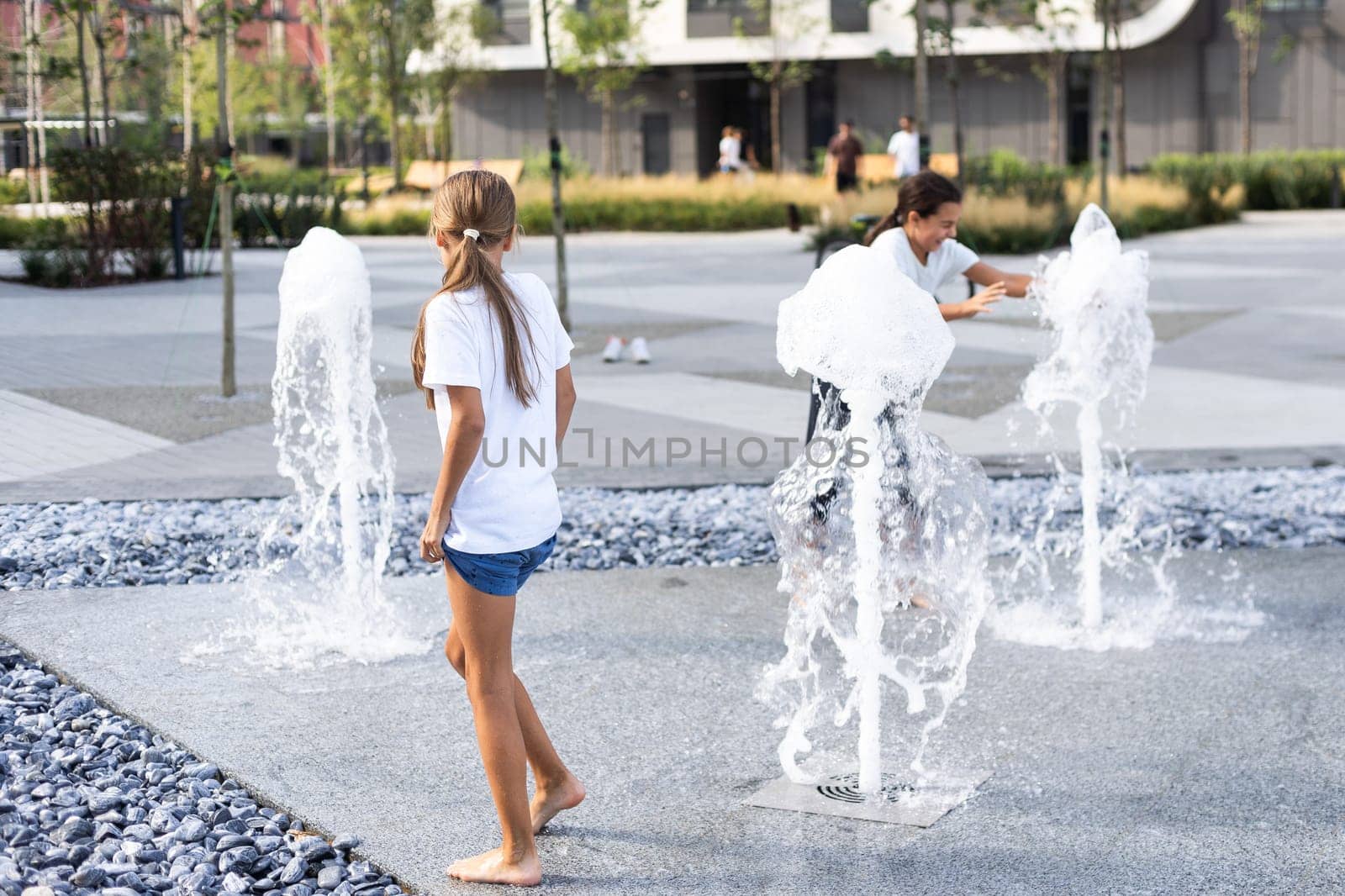 Cheerful young teen girl in city fountain, girl in wet clothes is having fun and enjoying the cool summer water, background city architecture. by Andelov13