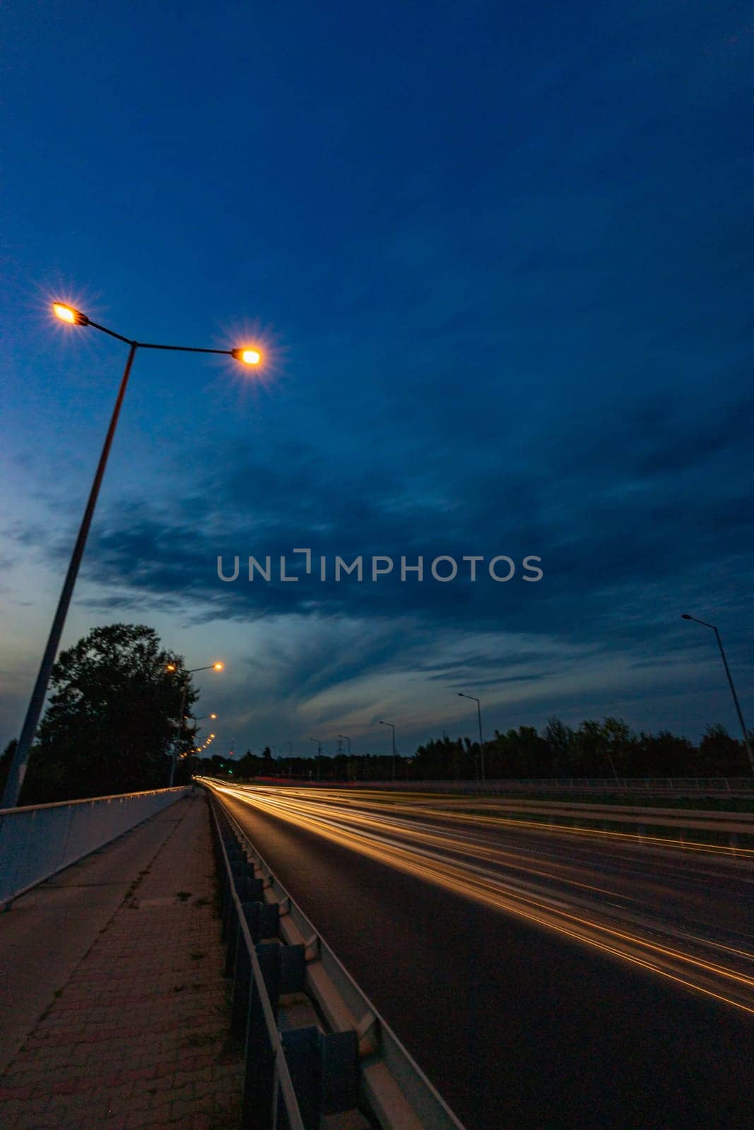 Long streaks of light made by riding cars on the road on the bridge at cloudy afternoon by Wierzchu