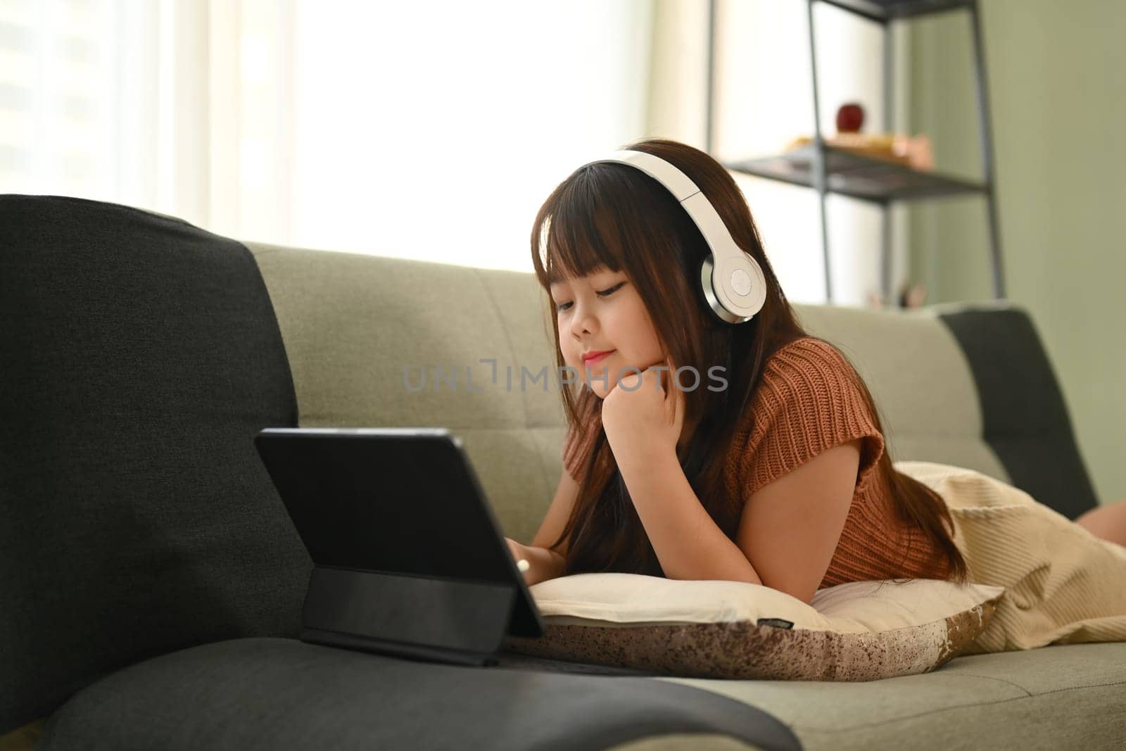 Little Asian girl in headphone lying on couch and watching video on digital tablet. by prathanchorruangsak