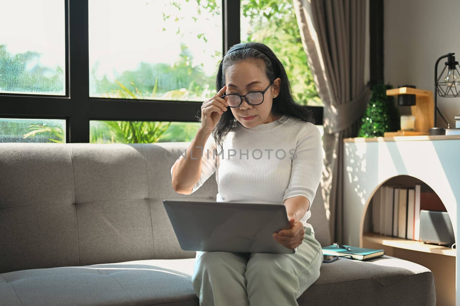 Middle age woman touching glasses and reading email on laptop while relaxing on the couch at home. by prathanchorruangsak