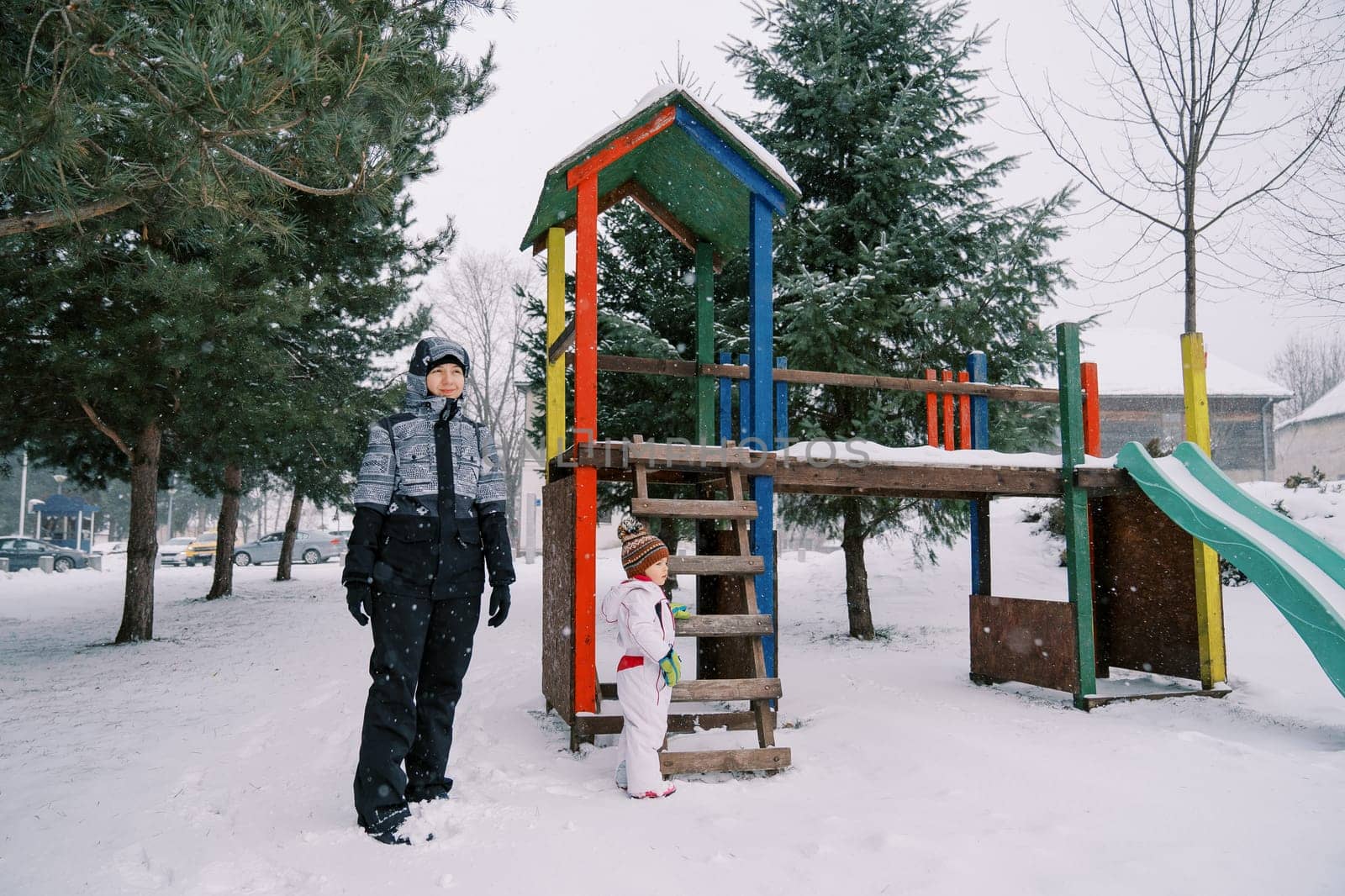 Mom and little girl stand near a snow-covered colorful slide among the trees by Nadtochiy