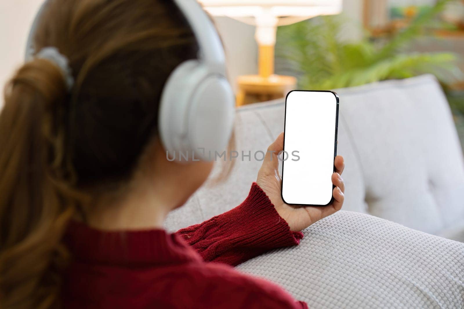 Mockup image of a woman holding mobile phone with blank desktop white screen and headphone while lying on a sofa at home.