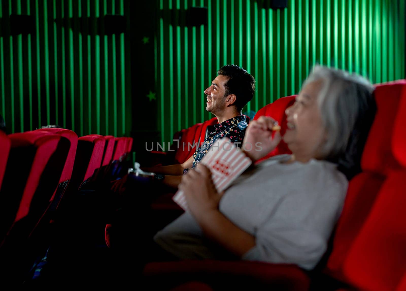 Caucasian man look happy with smiling during watch movie in cinema theater and senior woman also happy with eat popcorn as foreground.