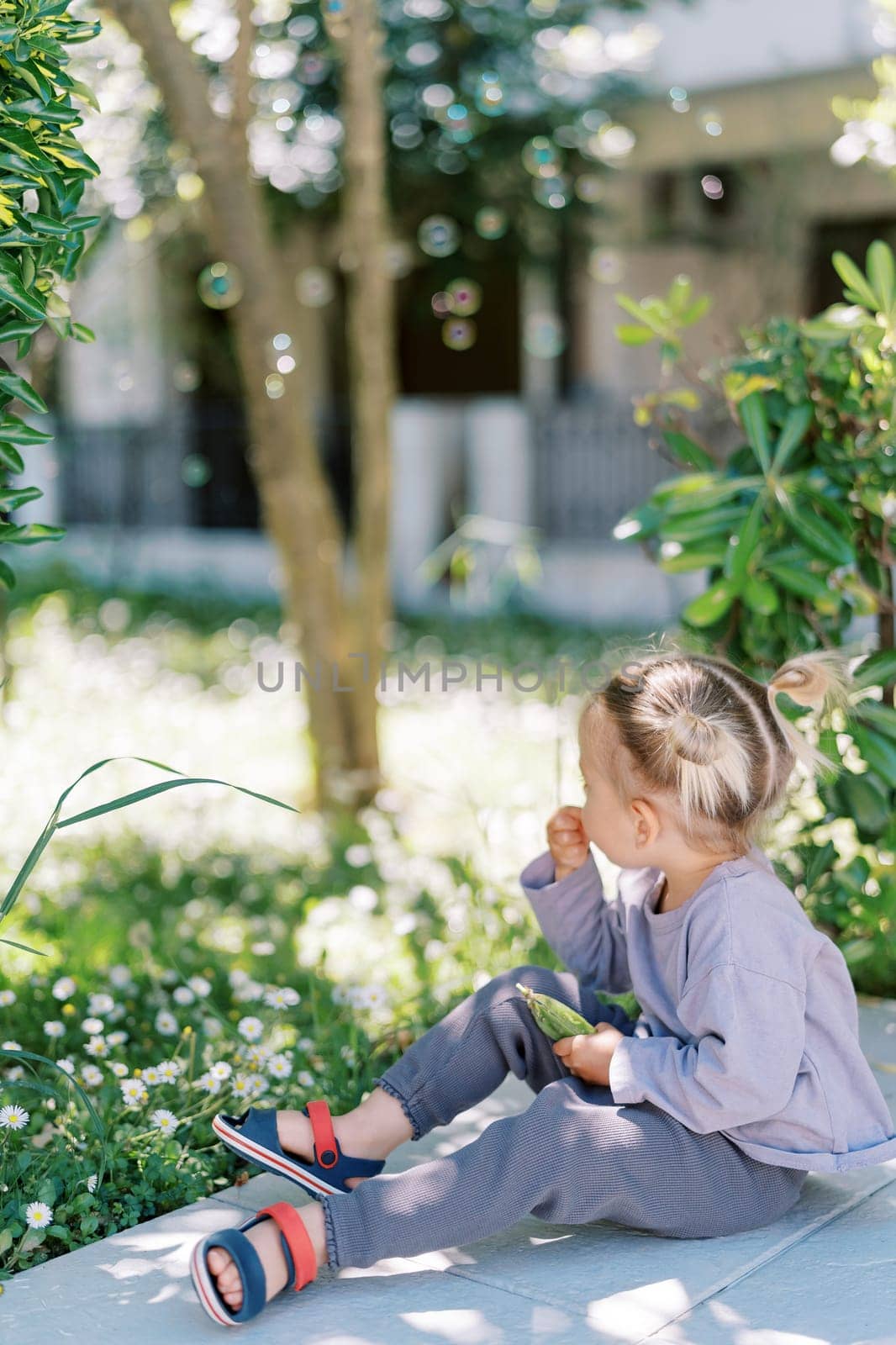 Little girl sits on a rug in the garden and eats green beans. Side view. High quality photo