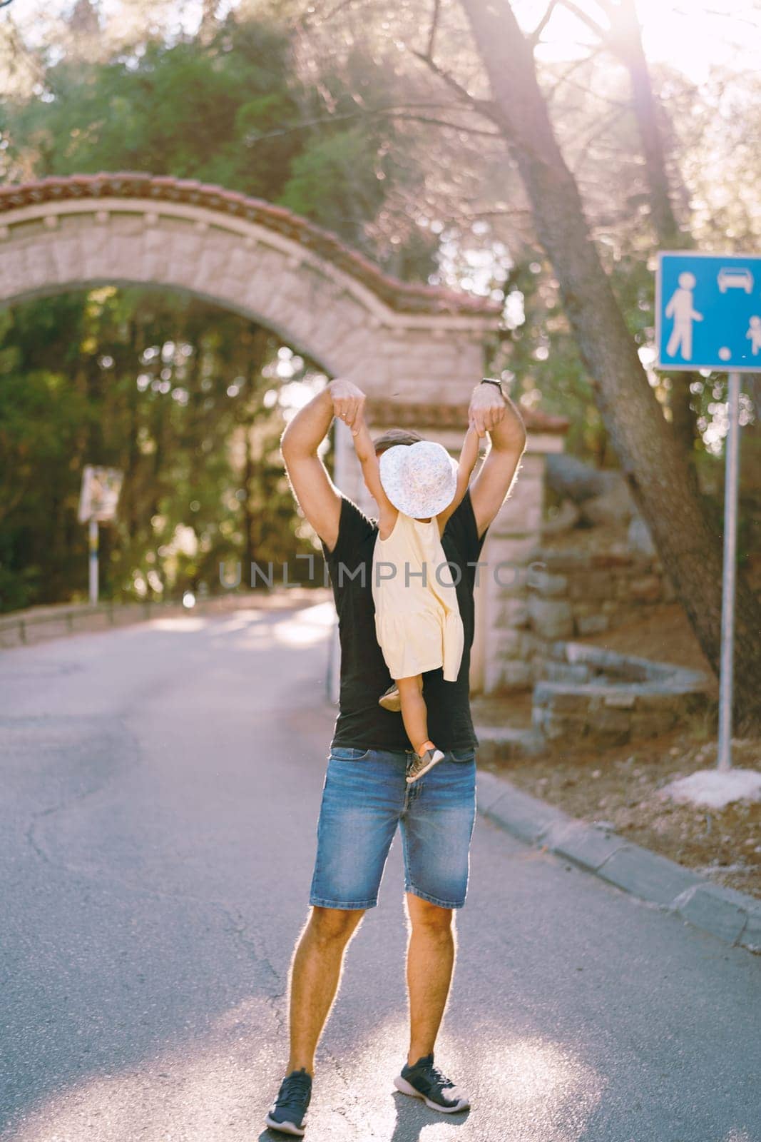 Dad lifts a little girl by the hands to the level of his face, standing on the road of the park. High quality photo