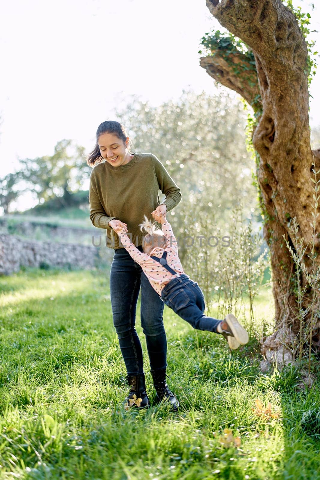 Smiling mother swings a little girl by the arms near a tree by Nadtochiy