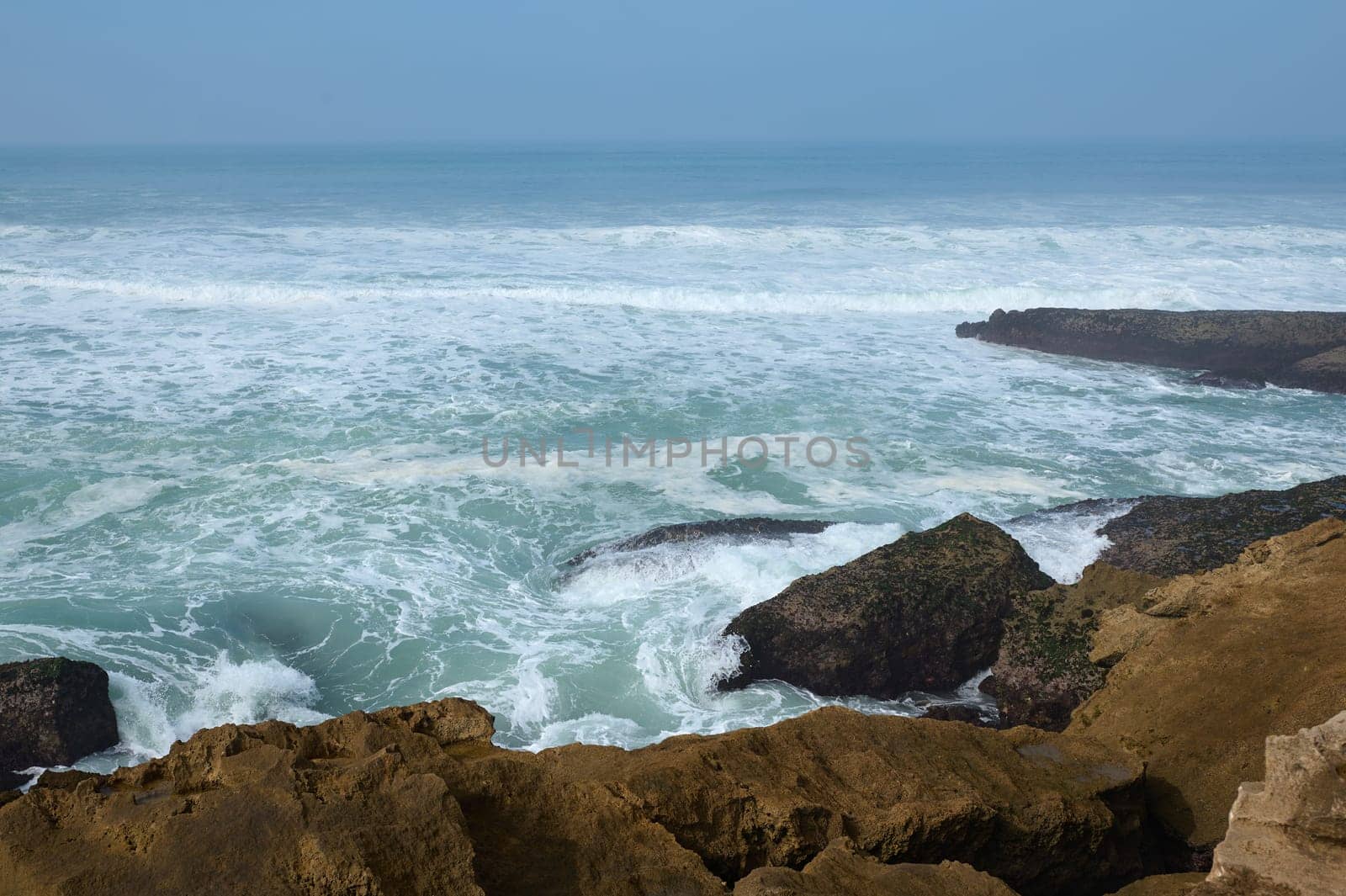 Beautiful waves making white sea foam while pounding on rocks and cliff of the Atlantic ocean shore. Clean clear blue sky on the horizon. Copy advertising space. Nature background. Landscape
