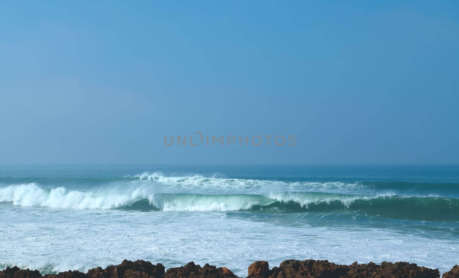 View of swirling waves in the Atlantic Ocean and blue clean clear sky on the horizon on sunny day. Seascape. Rocks and headland on the foreground. Nature background. Seashore. Copy space for ads