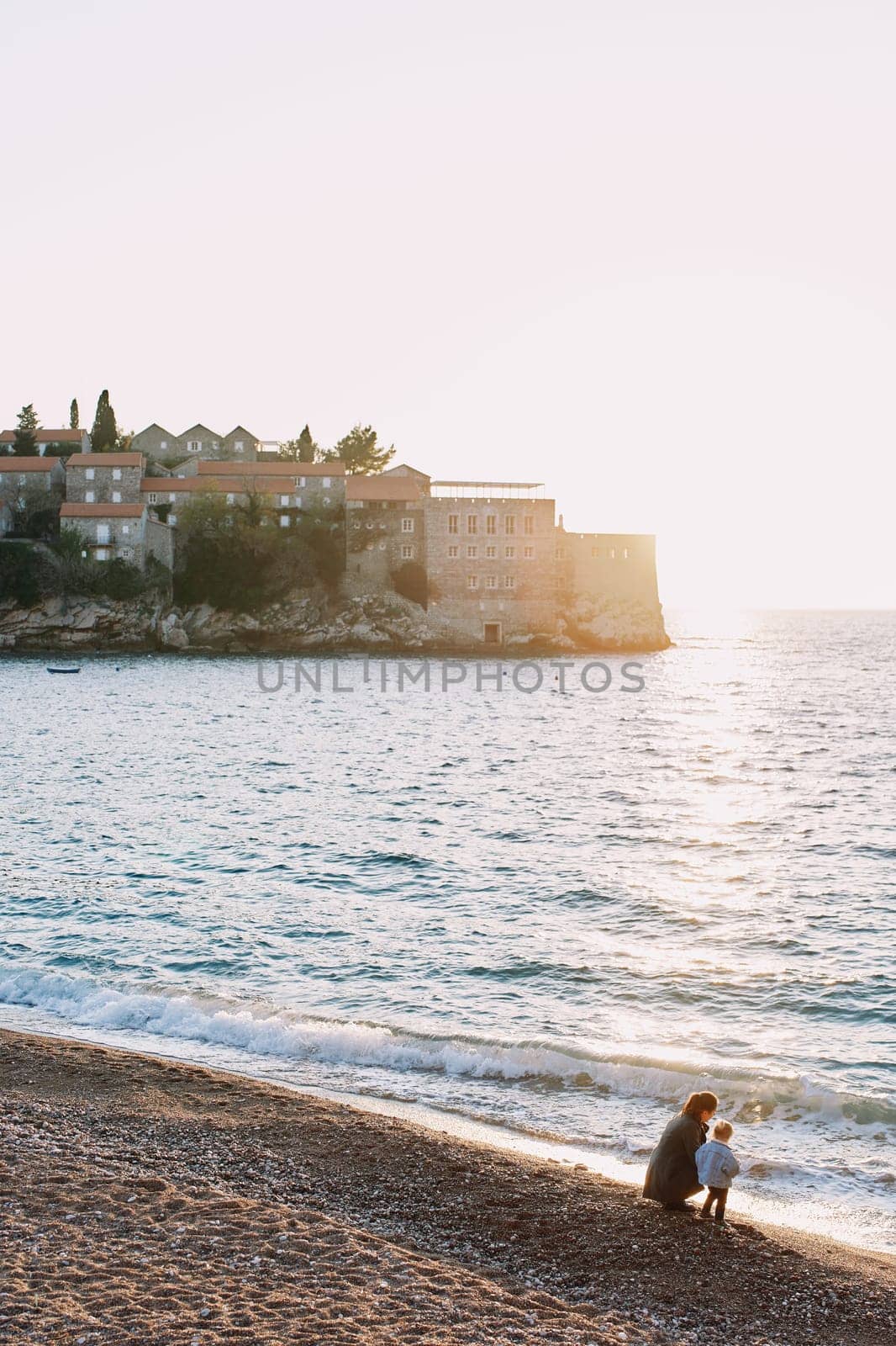 Mom and a little girl are sitting on the shore and looking at the sea, near the island of Sveti Stefan. Montenegro. High quality photo