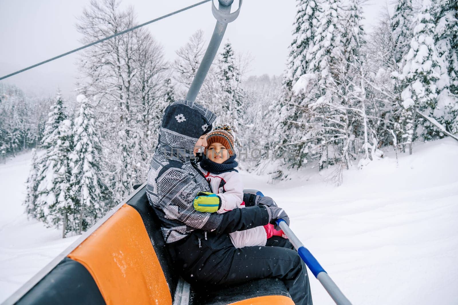 Smiling mother with a little girl in her arms rides a chairlift up a snowy hill by Nadtochiy