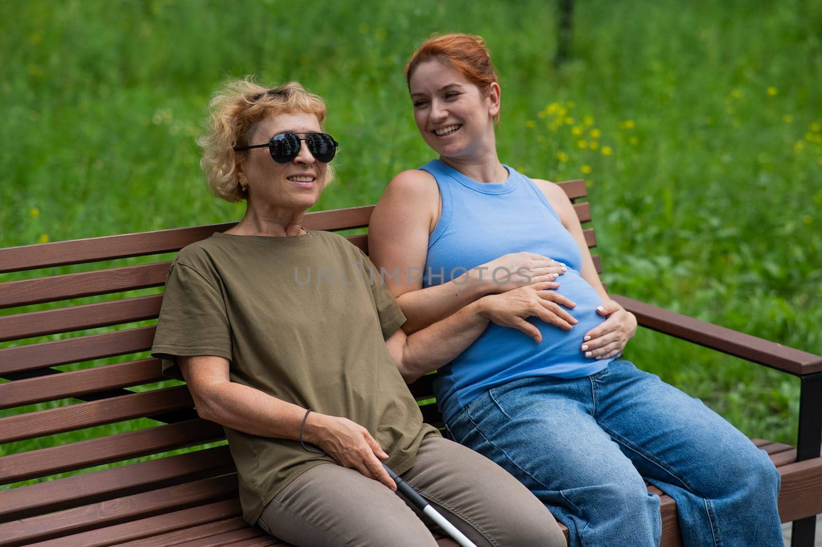An elderly blind woman holds her pregnant daughter by the belly while sitting on a bench in the park