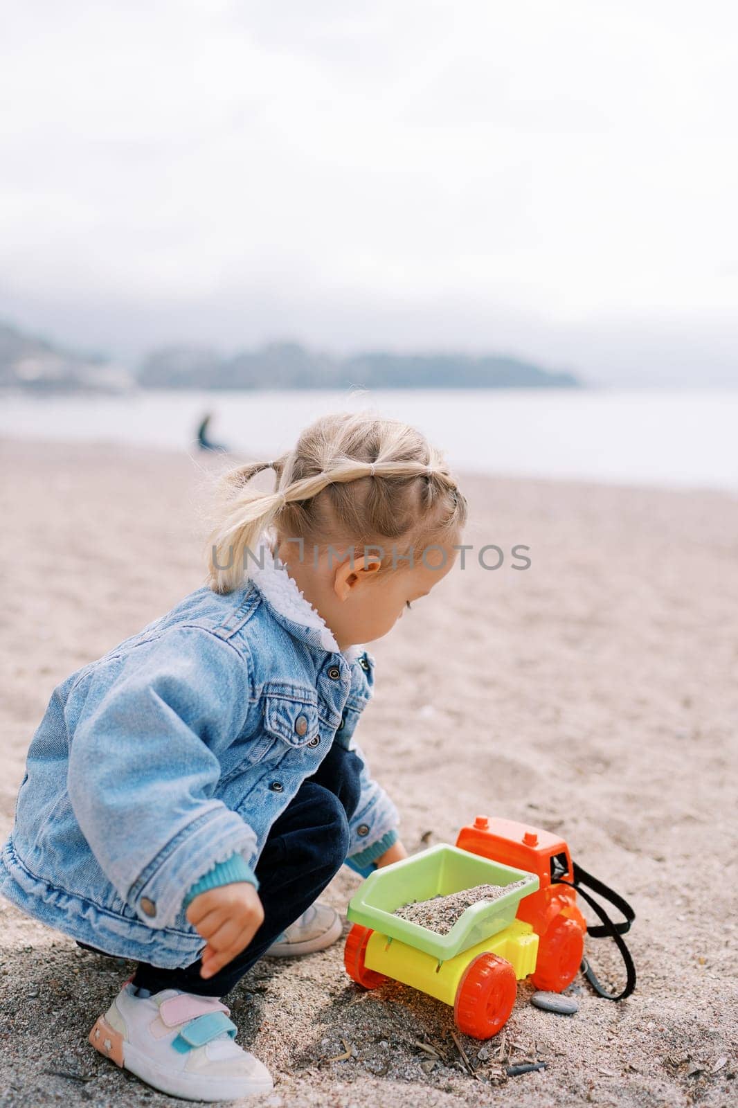 Little girl squats on the beach and pours sand into a toy car with her hands by Nadtochiy