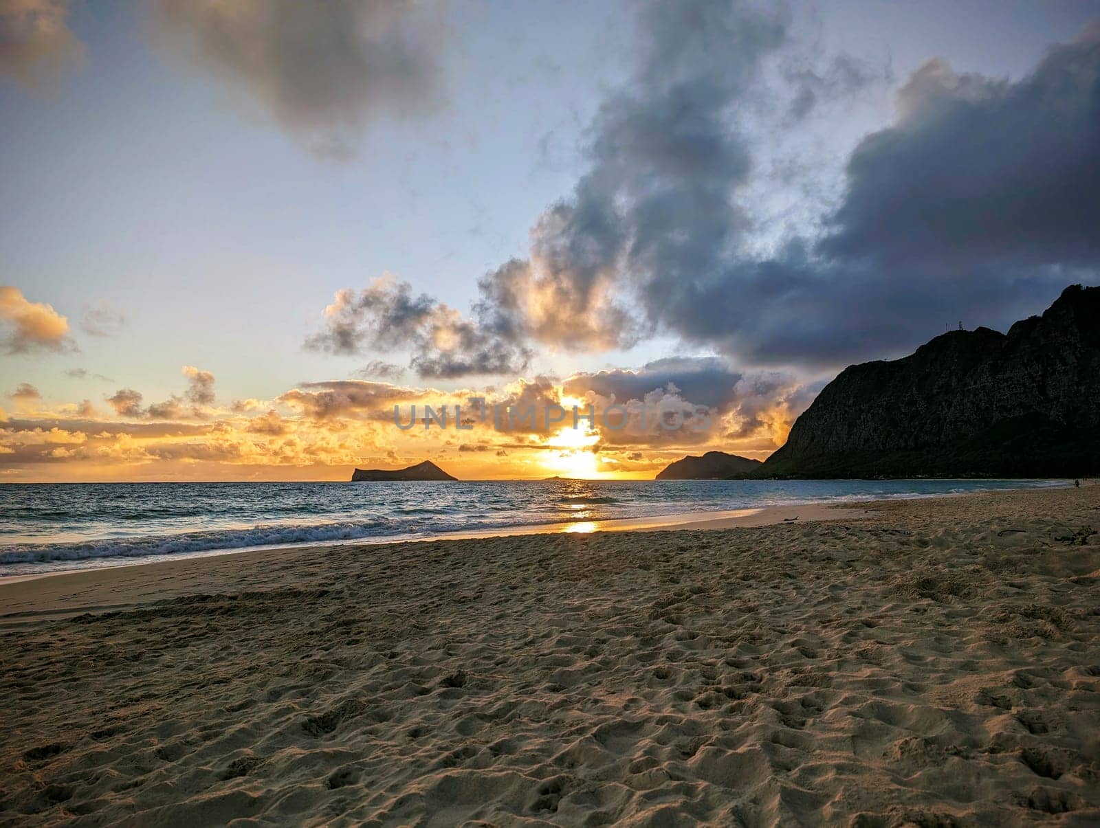 Sunrise on Waimanalo Beach, a long and sandy beach on the eastern coast of Oʻahu, Hawaii. The photo was taken from the perspective of someone standing on the beach, looking at the orange sun rising over the calm ocean. The sky is a mix of orange, pink, and blue colors, and there are a few clouds in the sky. 