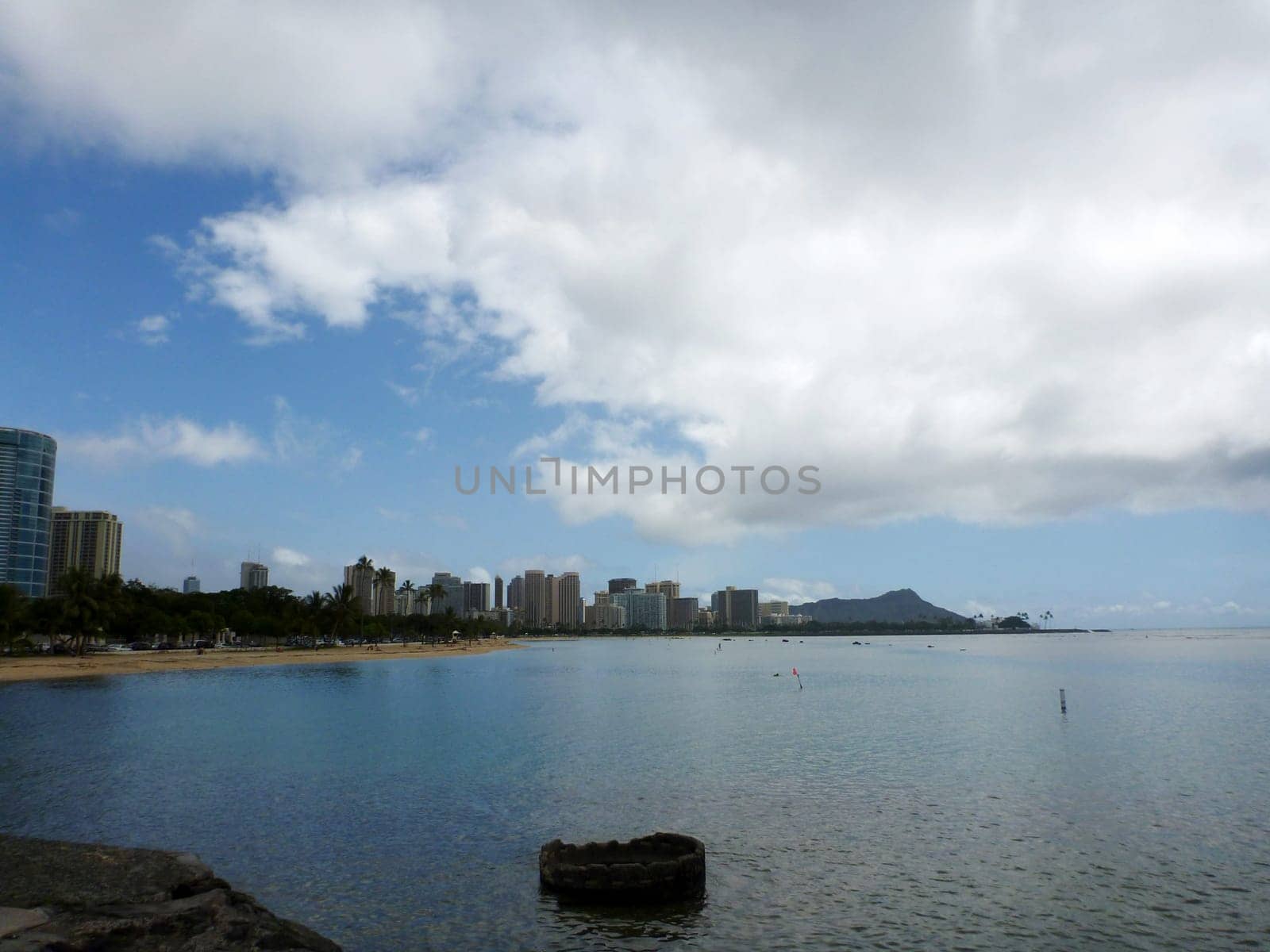 Stunning view of Honolulu’s skyline and the iconic Diamond Head crater from Ala Moana Beach Park, a popular recreational area in Hawaii. The photo was taken from the rock wall near Kewalo Basin, a harbor for fishing and recreational boats. The ocean is clear and blue, and the sky is partly cloudy.