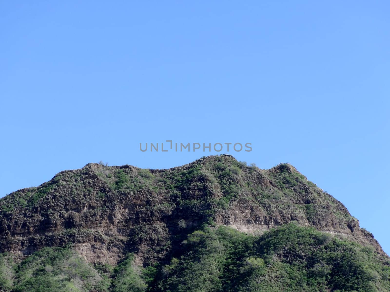 Green crater rim of Diamond Head, a volcanic tuff cone on the island of Oʻahu, Hawaii. The crater is a natural wonder that was formed by a volcanic eruption about 300,000 years ago. The crater is now a state monument and a popular hiking destination. The photo was taken from a distance, so the crater appears small in the frame, but it is actually 3,520 feet in diameter and 761 feet in height. The sky is a clear blue, and the crater contrasts with the surrounding landscape.