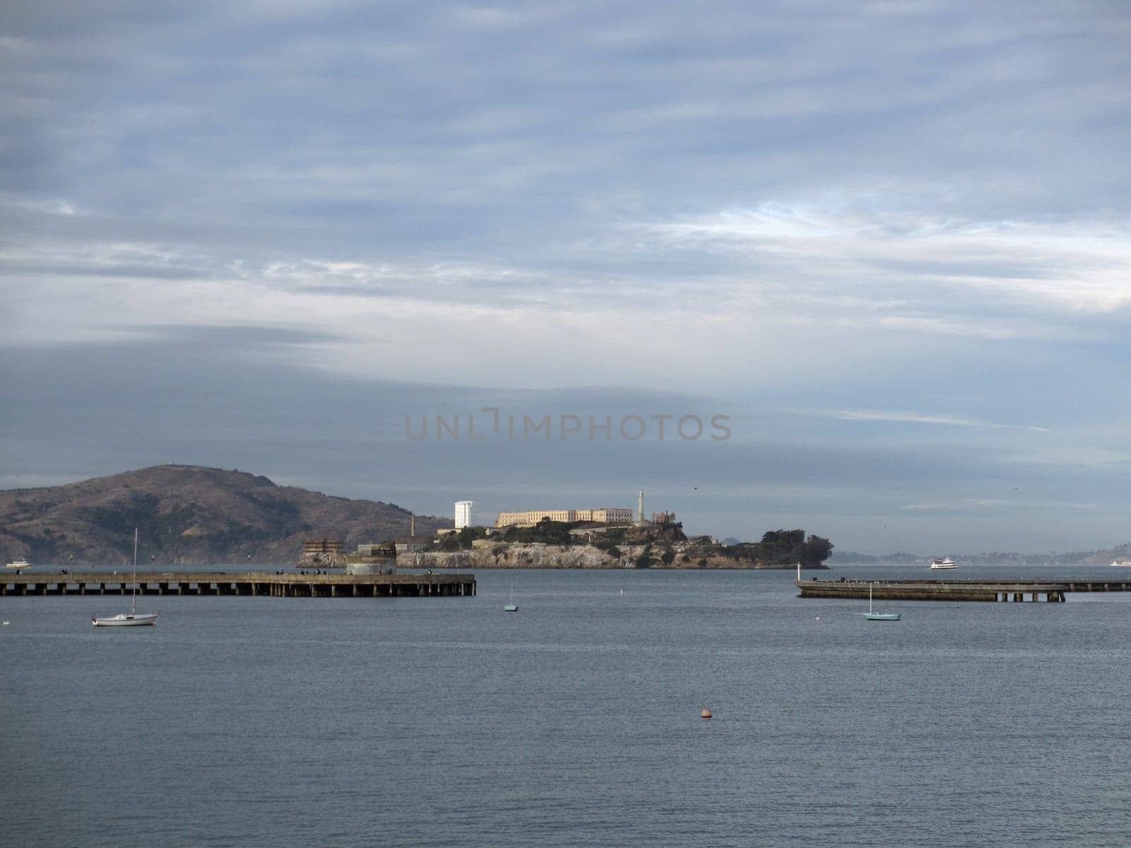 San Francisco - December 28, 2011: Long pier extending into the bay, leading towards Alcatraz Island under a cloudy sky. The photo shows several small boats scattered across the calm water, adding to the tranquil atmosphere. The photo is taken from a low angle, looking up at the pier and the island.