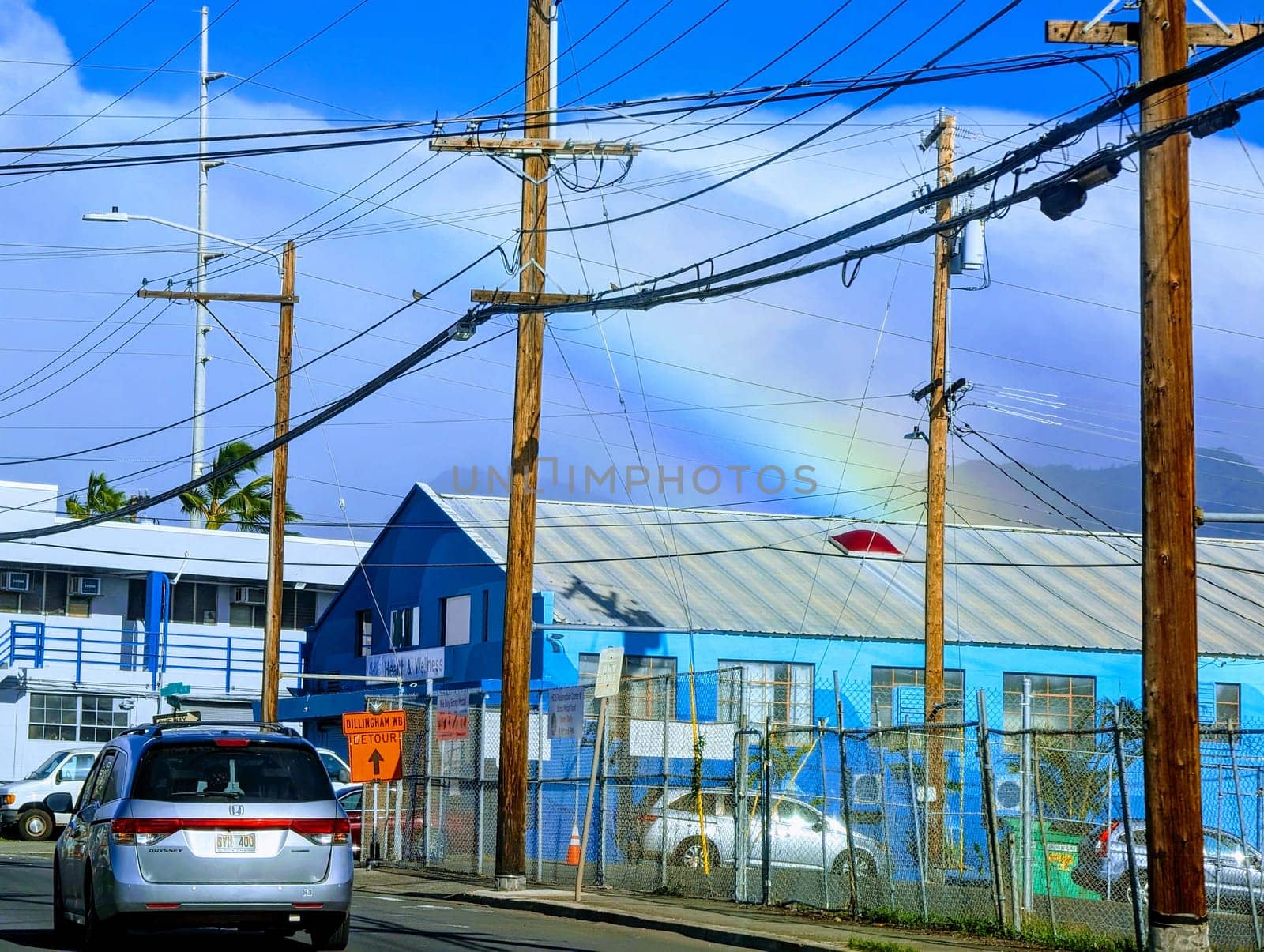 Honolulu - December 28, 2023: Street view in Kalihi, with a focus on utility poles, wires, a car, buildings, and a rainbow in the sky. The photo shows the intricate network of wires and the silver car parked next to a chain-link fence. The photo also shows the blue buildings with red roofs and the mountains in the background.