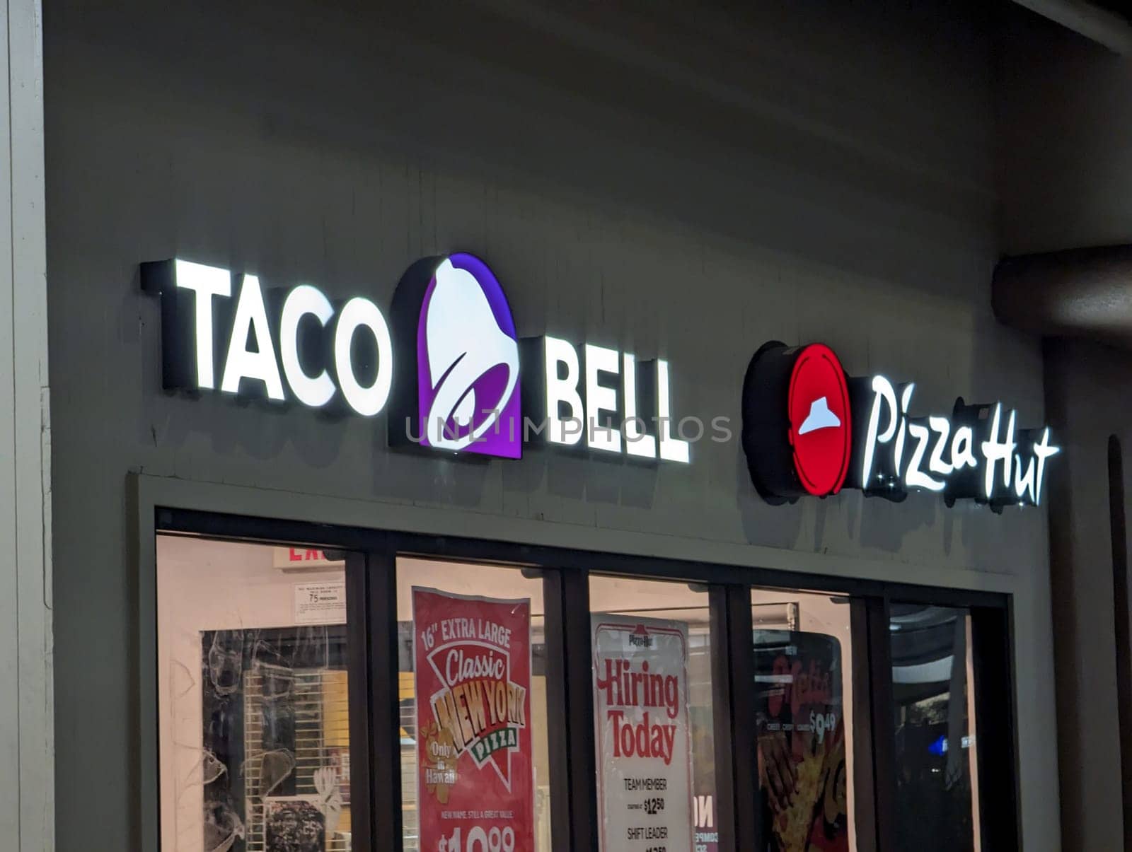 Honolulu - December 6, 2022: Taco Bell and Pizza Hut storefront in a strip mall at night. The storefront has a black awning with white lettering and logos. The windows display advertisements and a hiring sign. The parking lot and other stores are visible in the background.