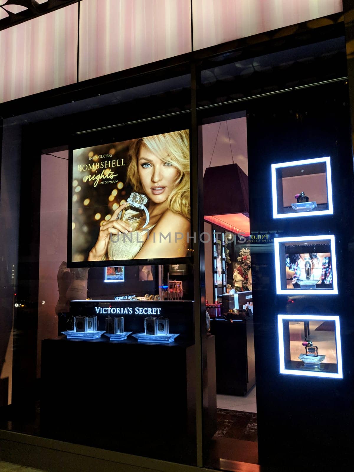 Waikiki - December 7, 2017:  Victoria’s Secret store window display at night in Waikiki, Hawaii. The display features a large poster of a woman wearing a black bra and holding a perfume bottle, and several smaller screens with images of lingerie and perfume. The window is framed in black and has a pink striped awning. 