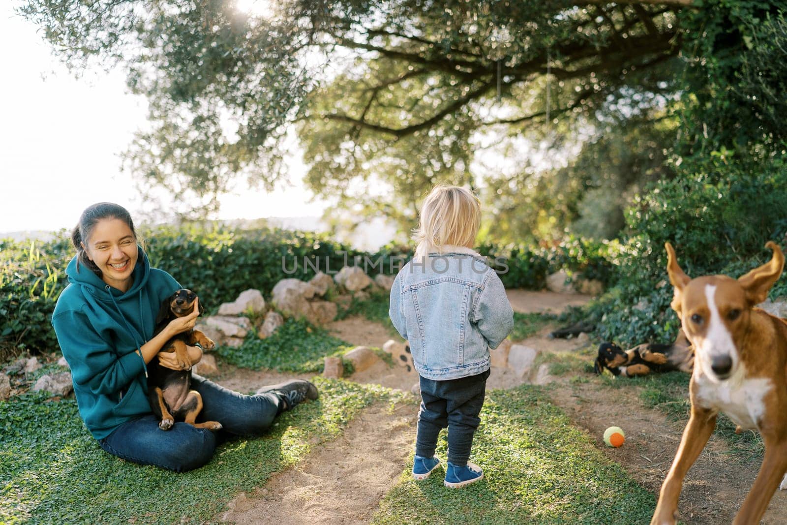 Little girl stands in front of a laughing mother sitting on the grass with a puppy in her arms. High quality photo