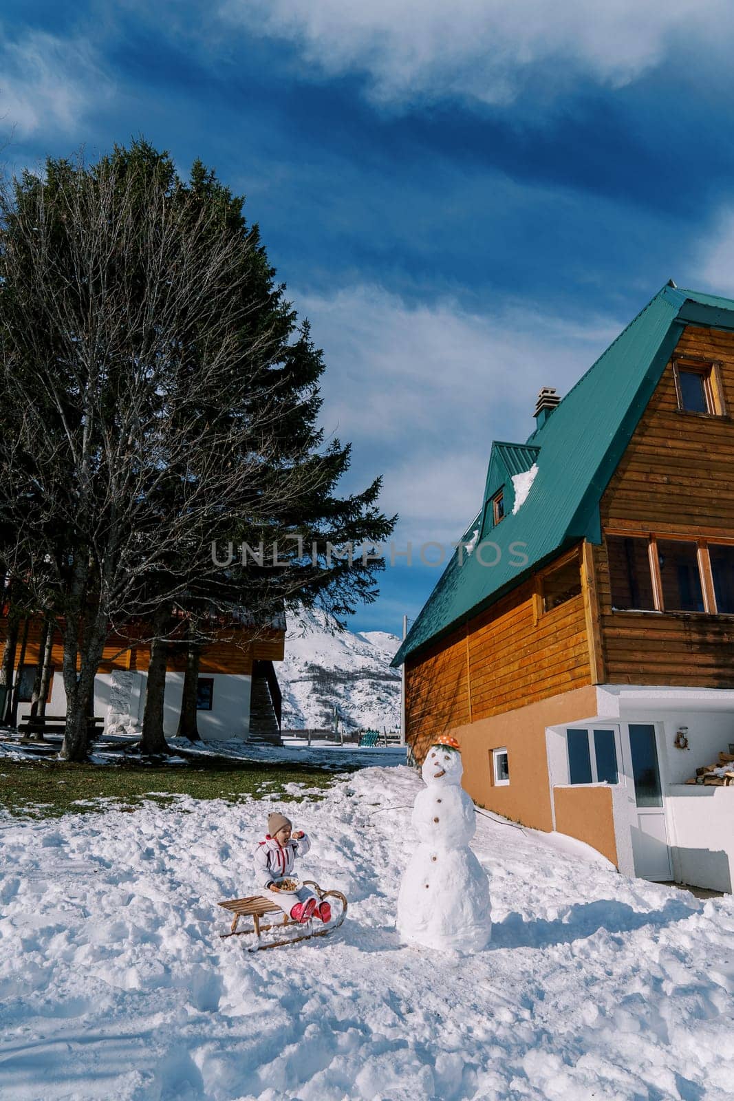 Little girl eats from a plate sitting on a sleigh near a snowman next to a wooden house. High quality photo