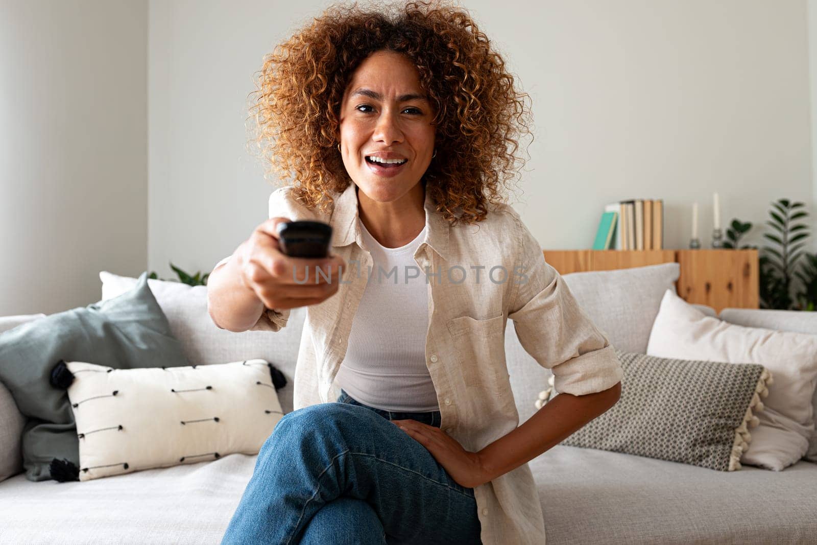 Happy African American woman watching TV sitting on the sofa using remote control to change channel looking at camera. by Hoverstock