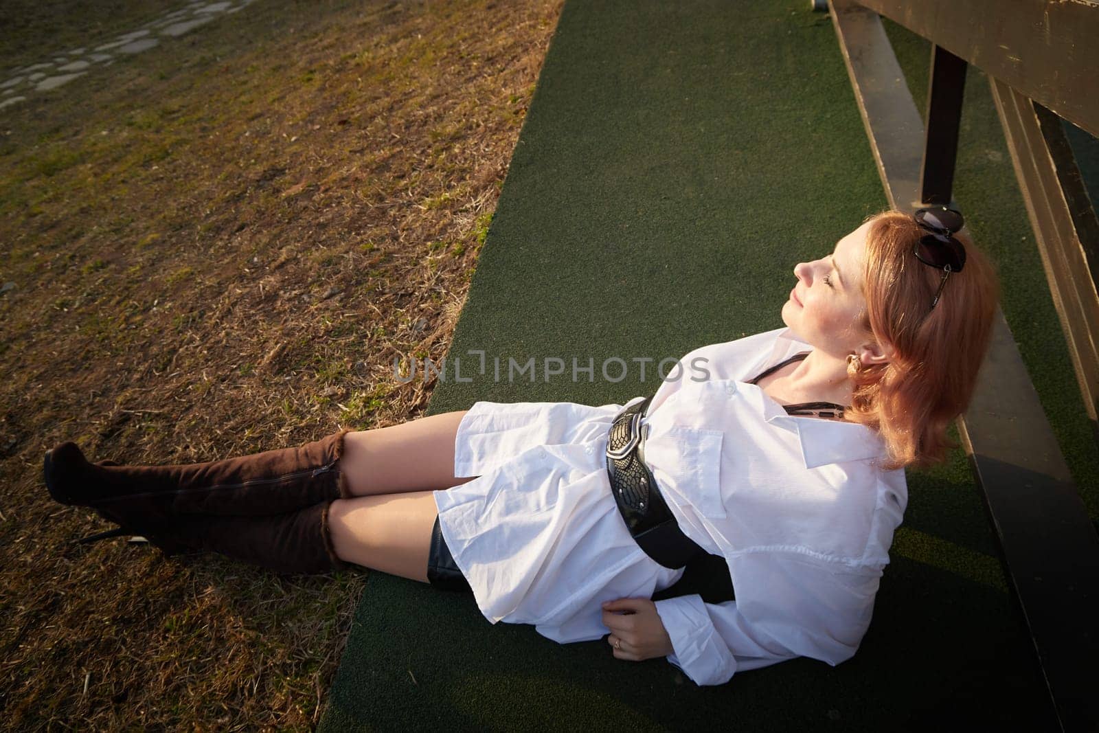 Beautiful girl with red hair in white shirt in open wooden pavillion in village or small town. Young slender woman and sky with clouds on background on autumn, spring or summer day