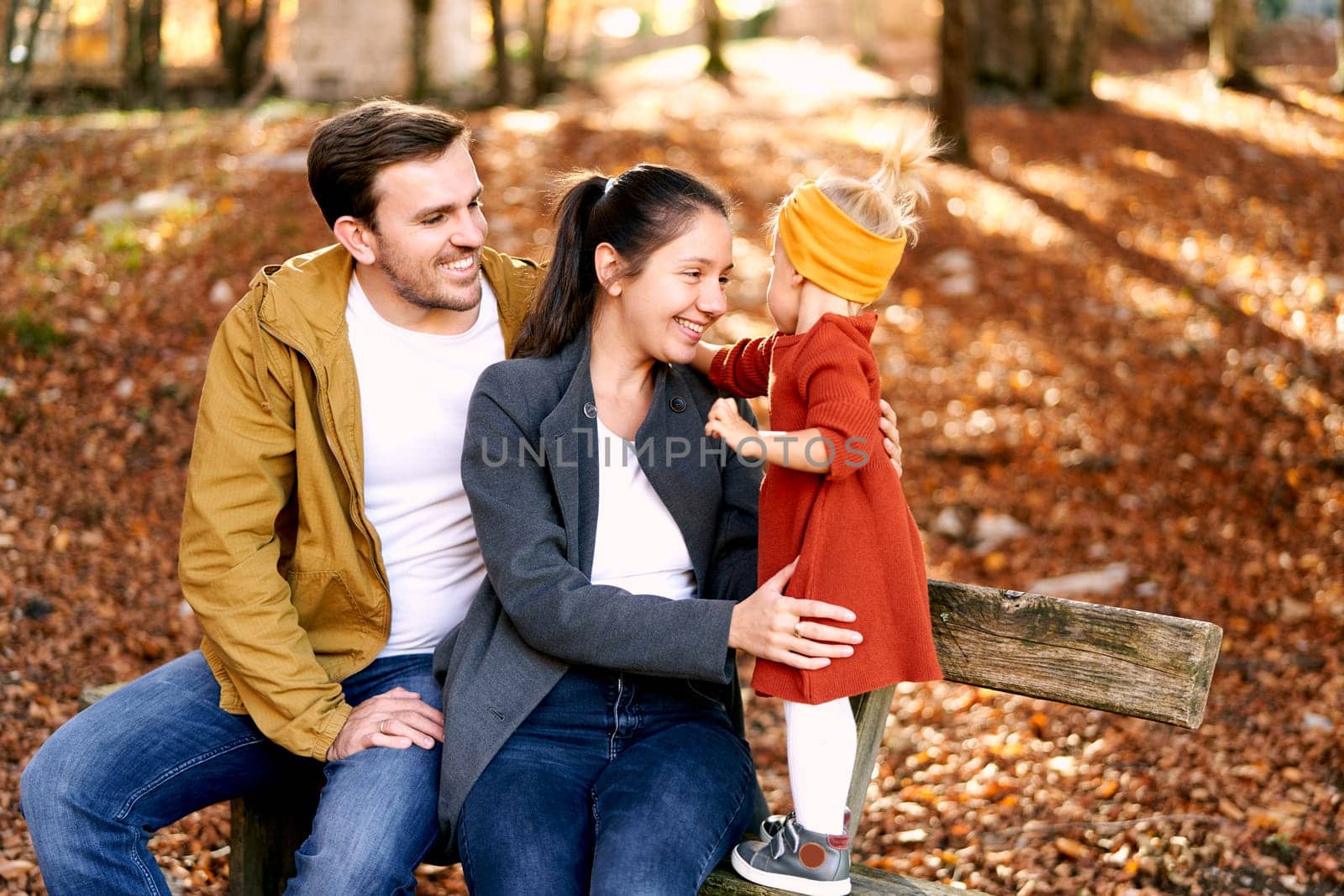 Little girl touches dad sitting with mom on bench in autumn forest. High quality photo