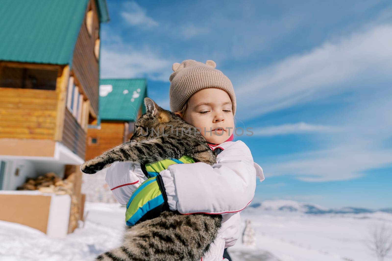 Little girl hugs a tabby cat while standing in the snow near a wooden cottage by Nadtochiy