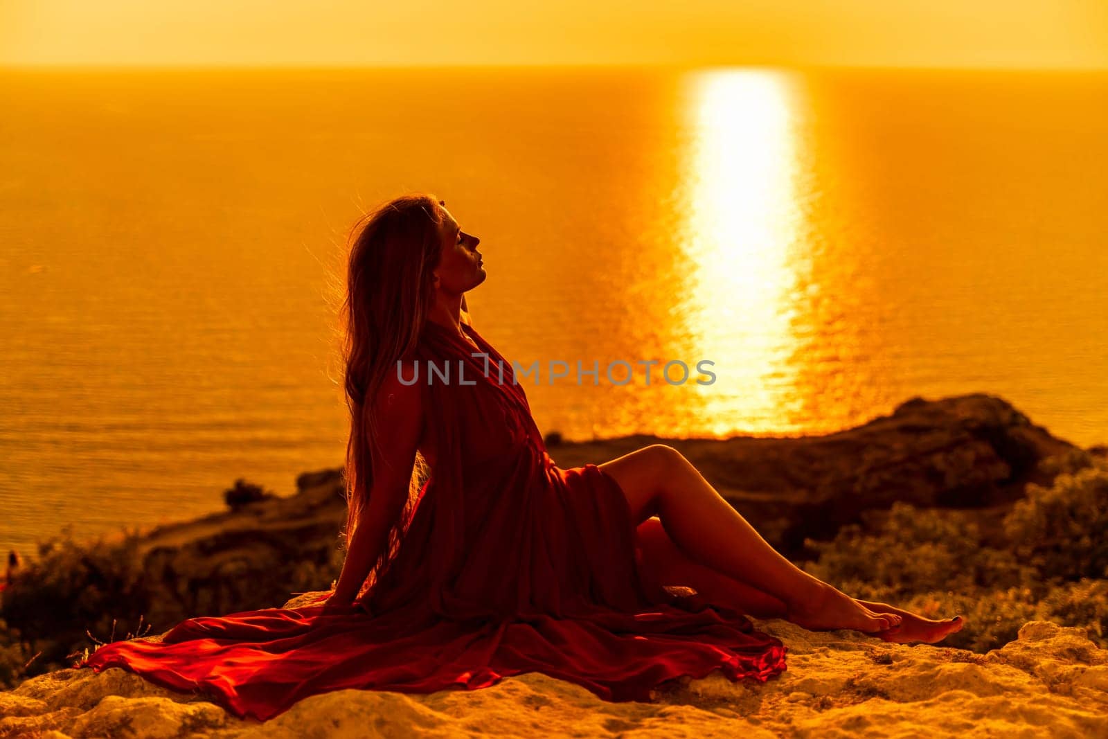 Woman sunset sea red dress, side view a happy beautiful sensual woman in a red long dress posing on a rock high above the sea on sunset