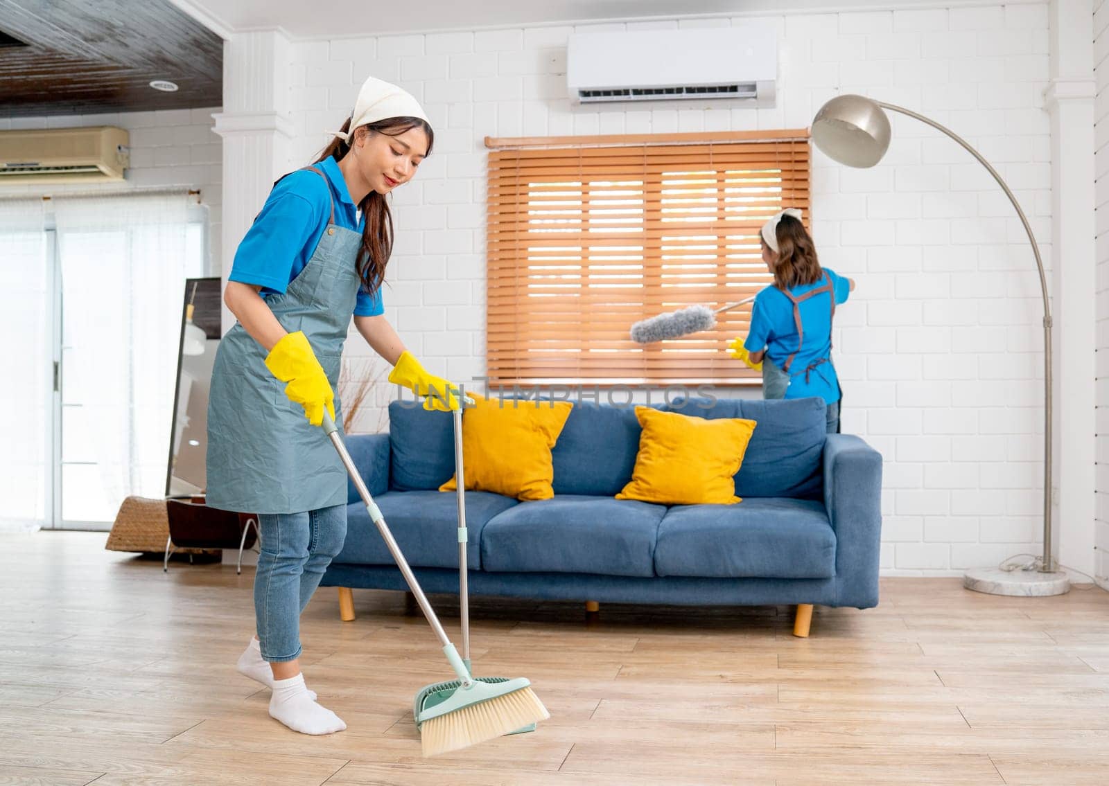 Wide shot of Asian housemaid or housekeeper team help to clean living room together with one use broom clean floor and other use feather duster for cleaning window.