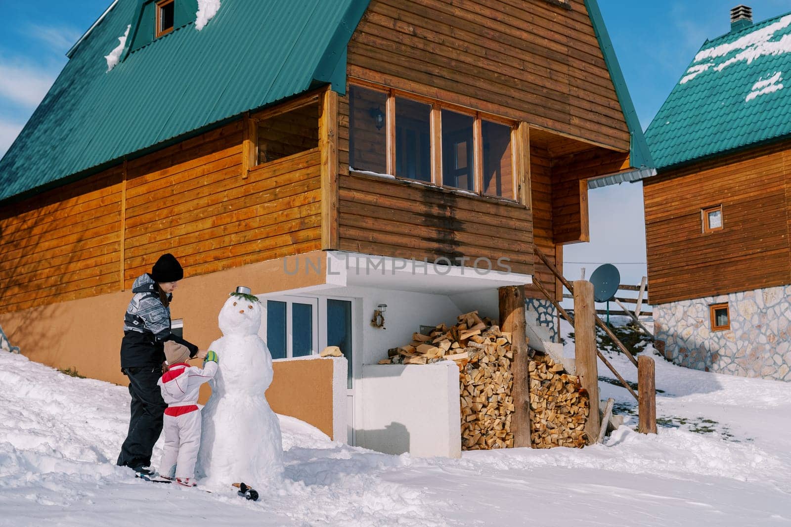 Mom and little girl decorate a snowman near a woodpile of a wooden cottage in the snow by Nadtochiy