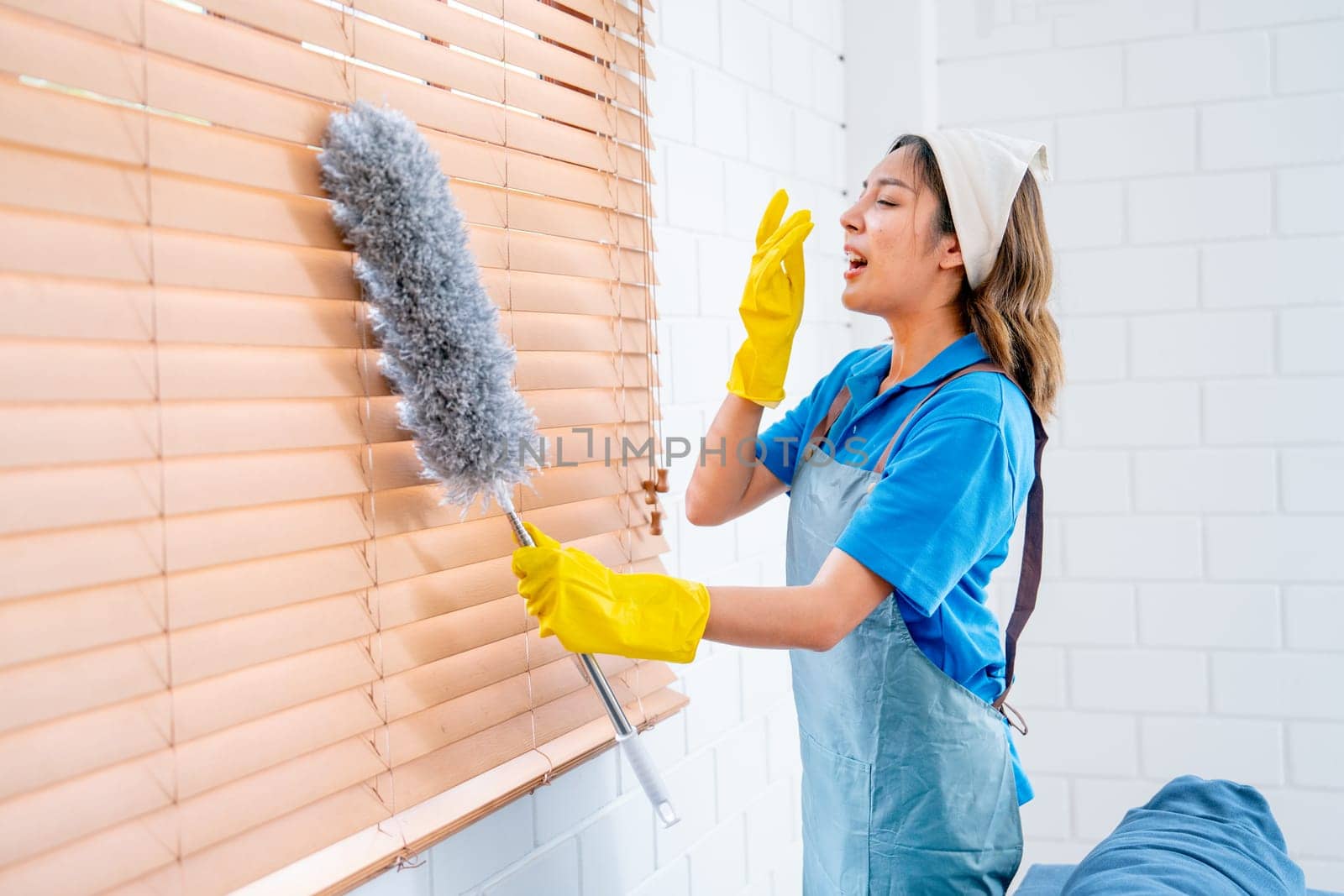 Asian housekeeper or housemaid action of sneeze during clean curtain or shade of windows with feather duster in living room with day light.