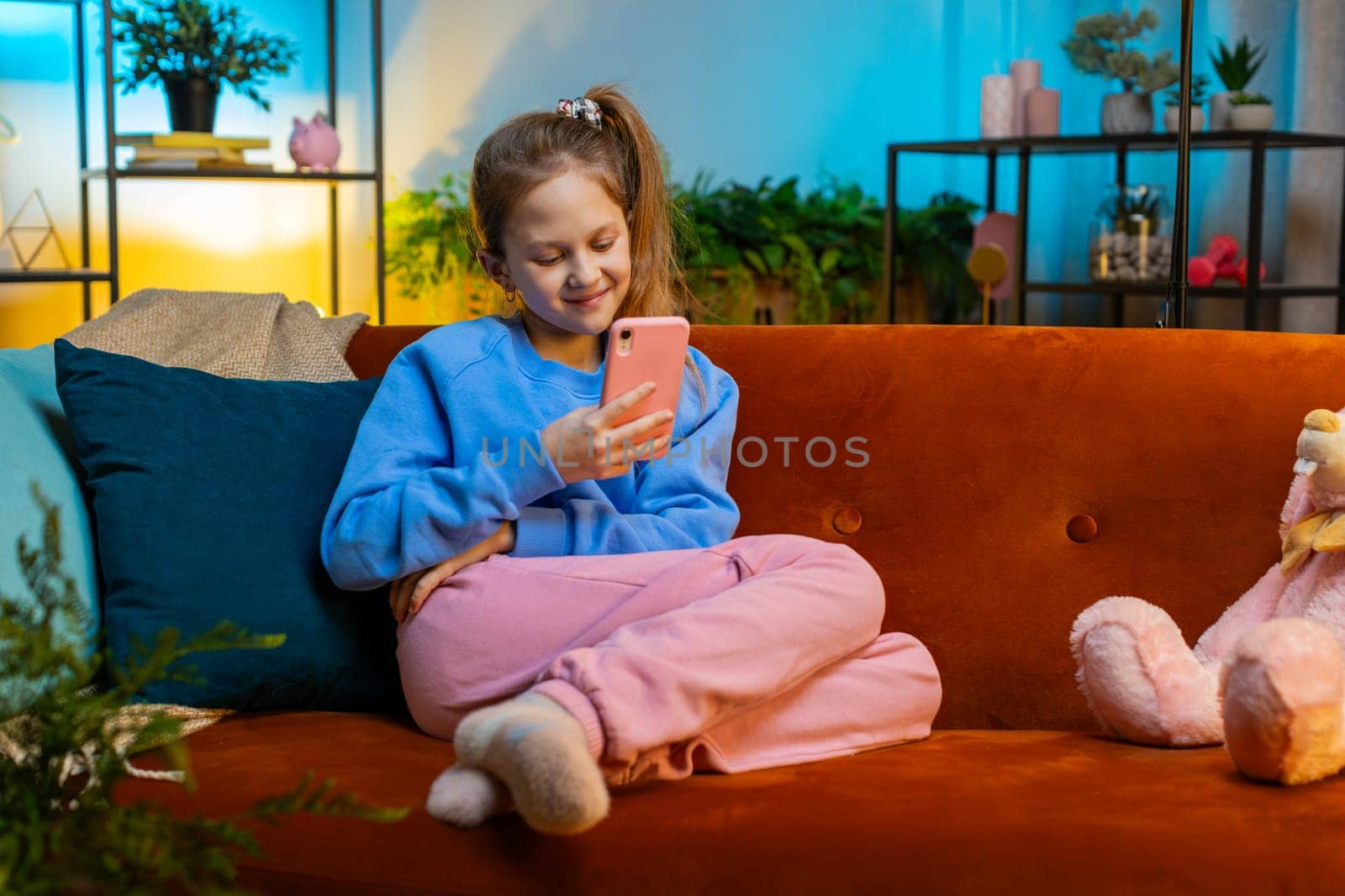 Young child girl texting share messages content on smartphone social media applications online, watching relax movie. Female teenager kid uses mobile phone at home in evening night room sits on sofa