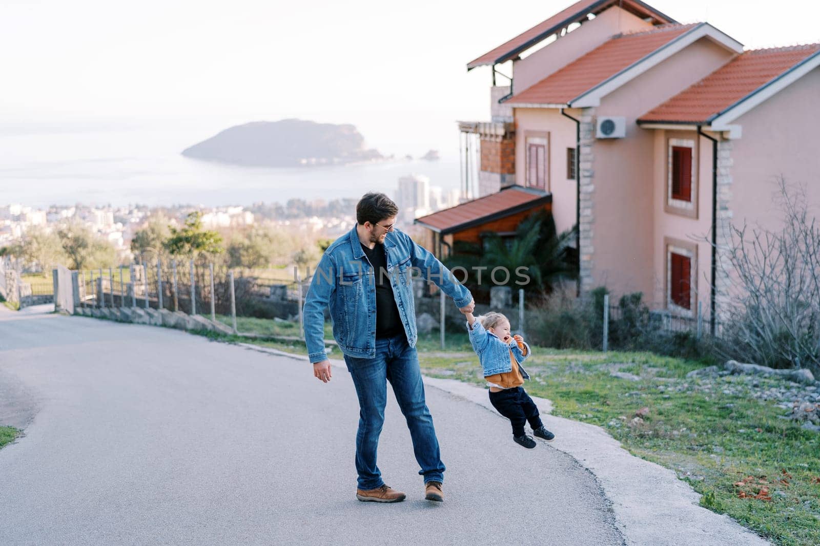 Dad is circling a little girl holding her hand on the road on a slope in a village above the sea. High quality photo