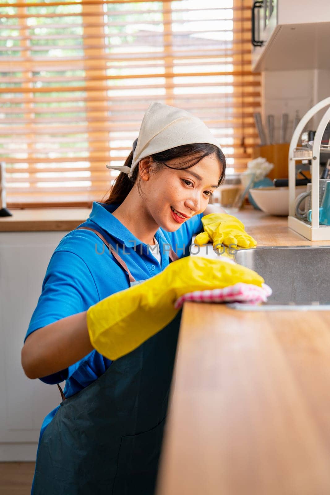 Vertical image of Asian housekeeper smile and sit with use towel to clean area in kitchen near sink with day light and she look happy to work in house.