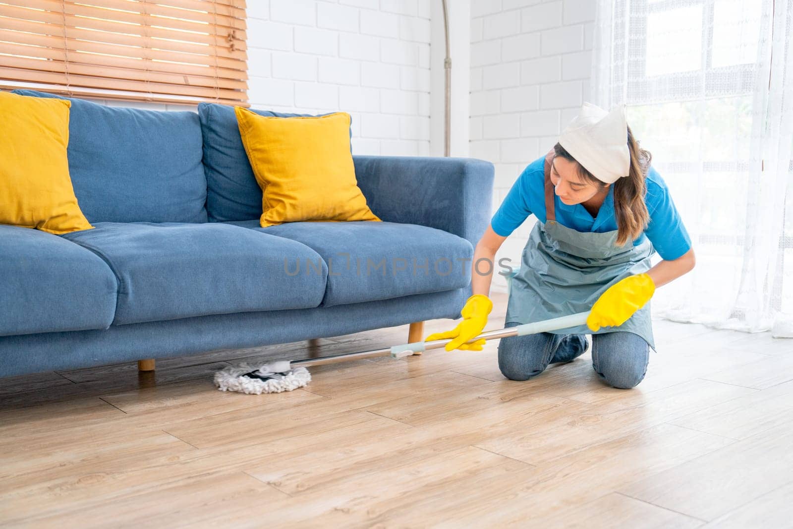 Asian housekeeper or housemaid woman use mop to clean under sofa in living room of the house with day light.