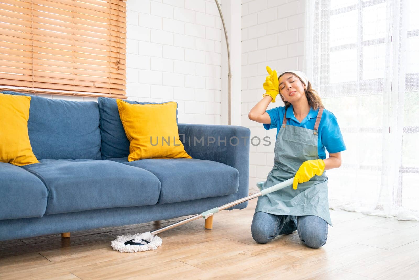 Asian housekeeper or housemaid woman action of tired after to use mop to clean under sofa in living room of the house with day light.