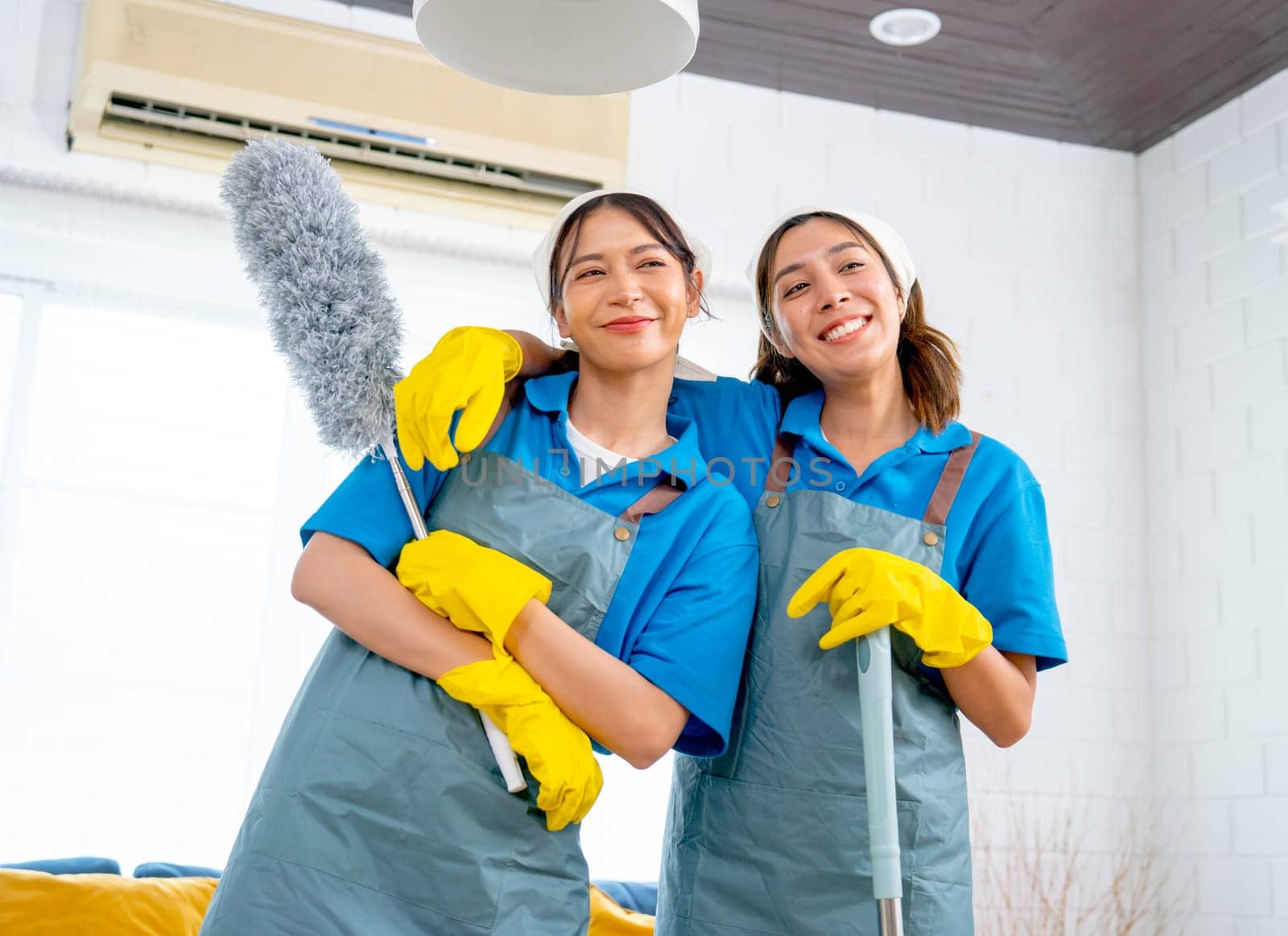 Portrait of Asian housekeeper or housemaid woman with hug together and stand with feather duster also look forward with happiness in living room of the house.