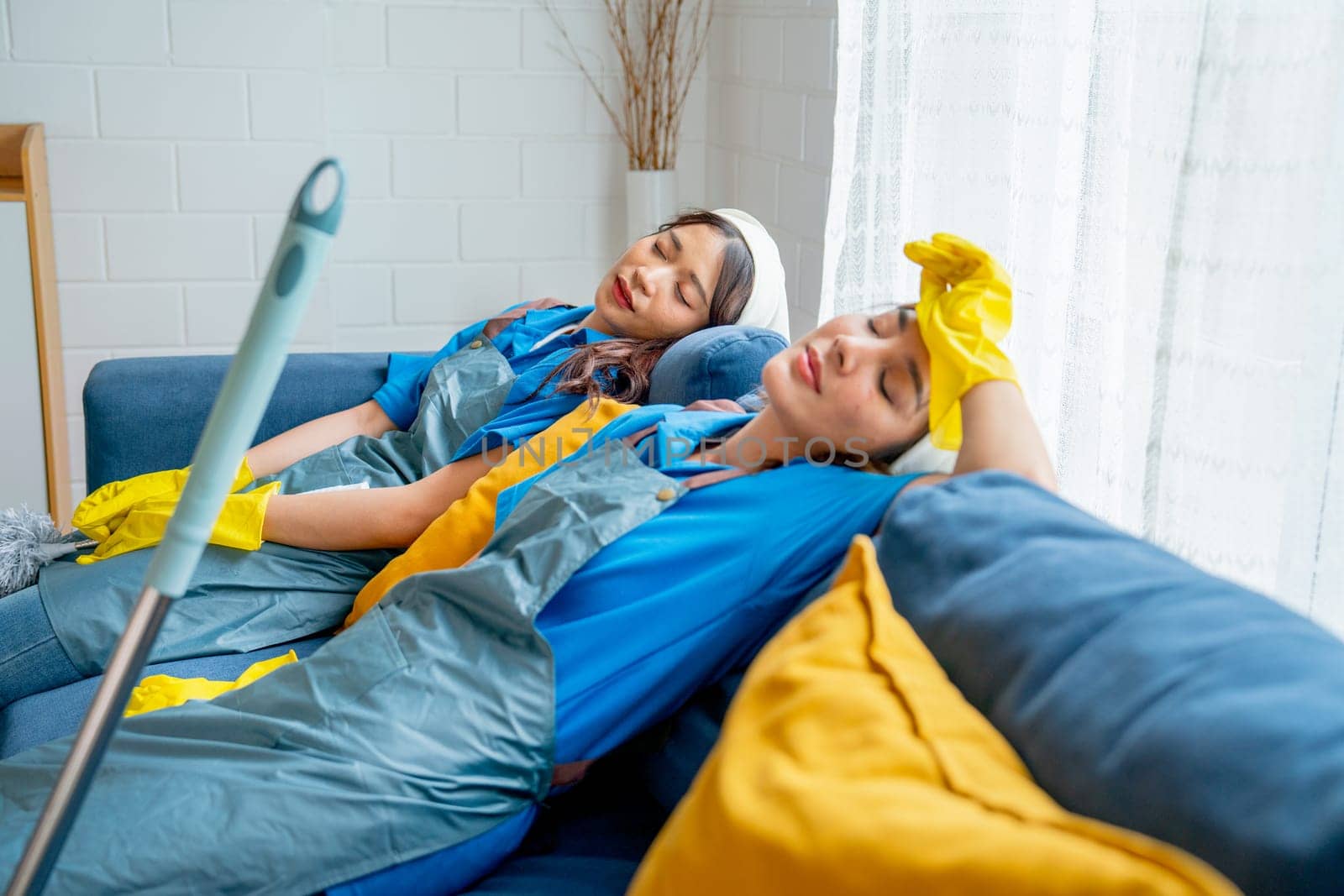 Two Asian housekeeper or housemaid women lie and sleep on sofa after feel tired from cleaning the house together with day light.