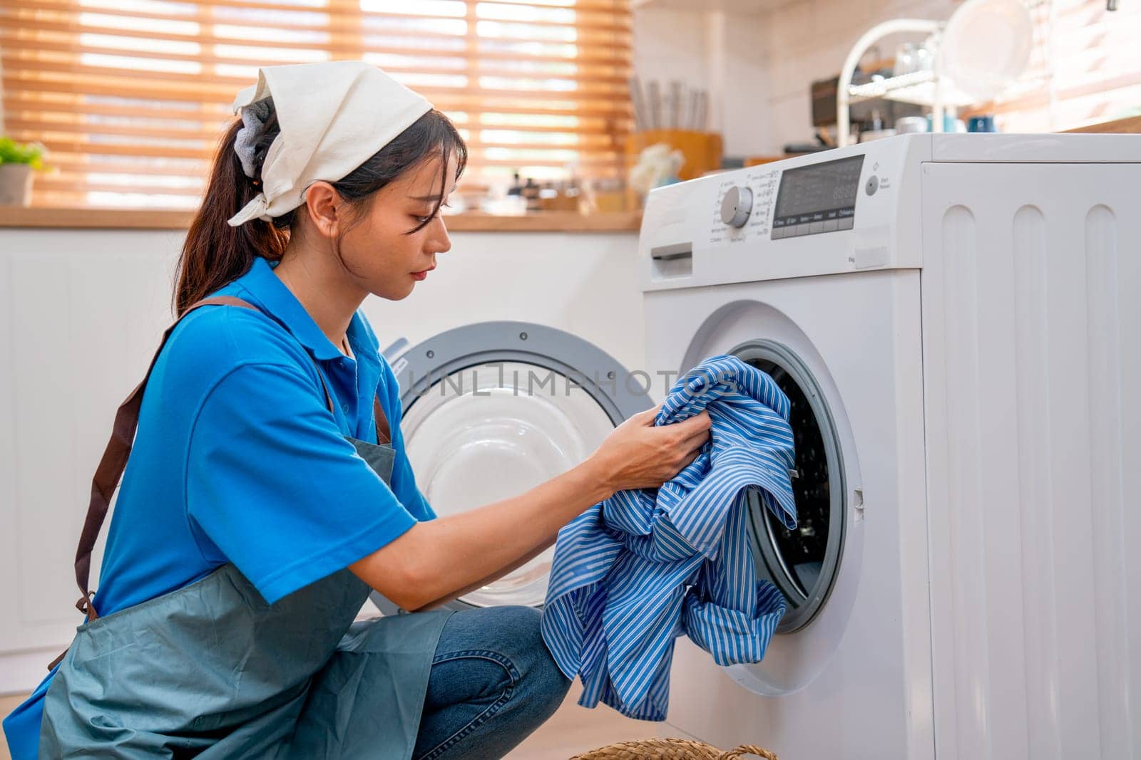 Asian housekeeper or housemaid put blue cloth into washing machine during work in customer house.