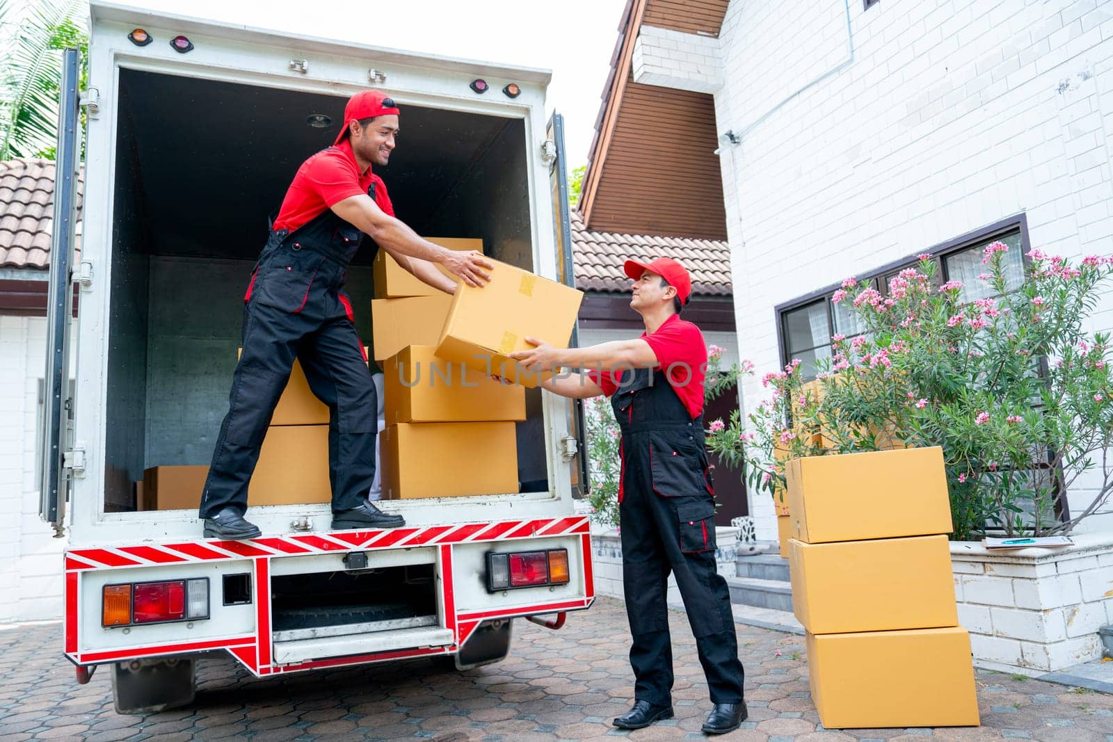 Wide shot of delivery man stand on truck and send parcel or box to his co-worker during transport the product to customer house. by nrradmin