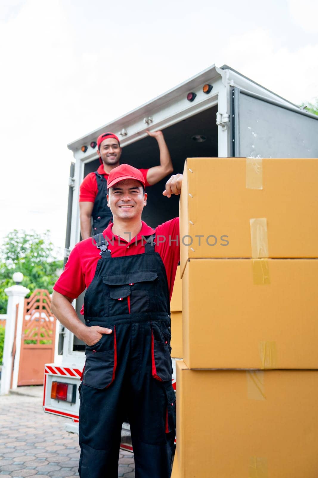 Vertical image and portrait of delivery man stand near stack of boxes and look at camera with smiling and his co-worker stand in the back on truck. by nrradmin
