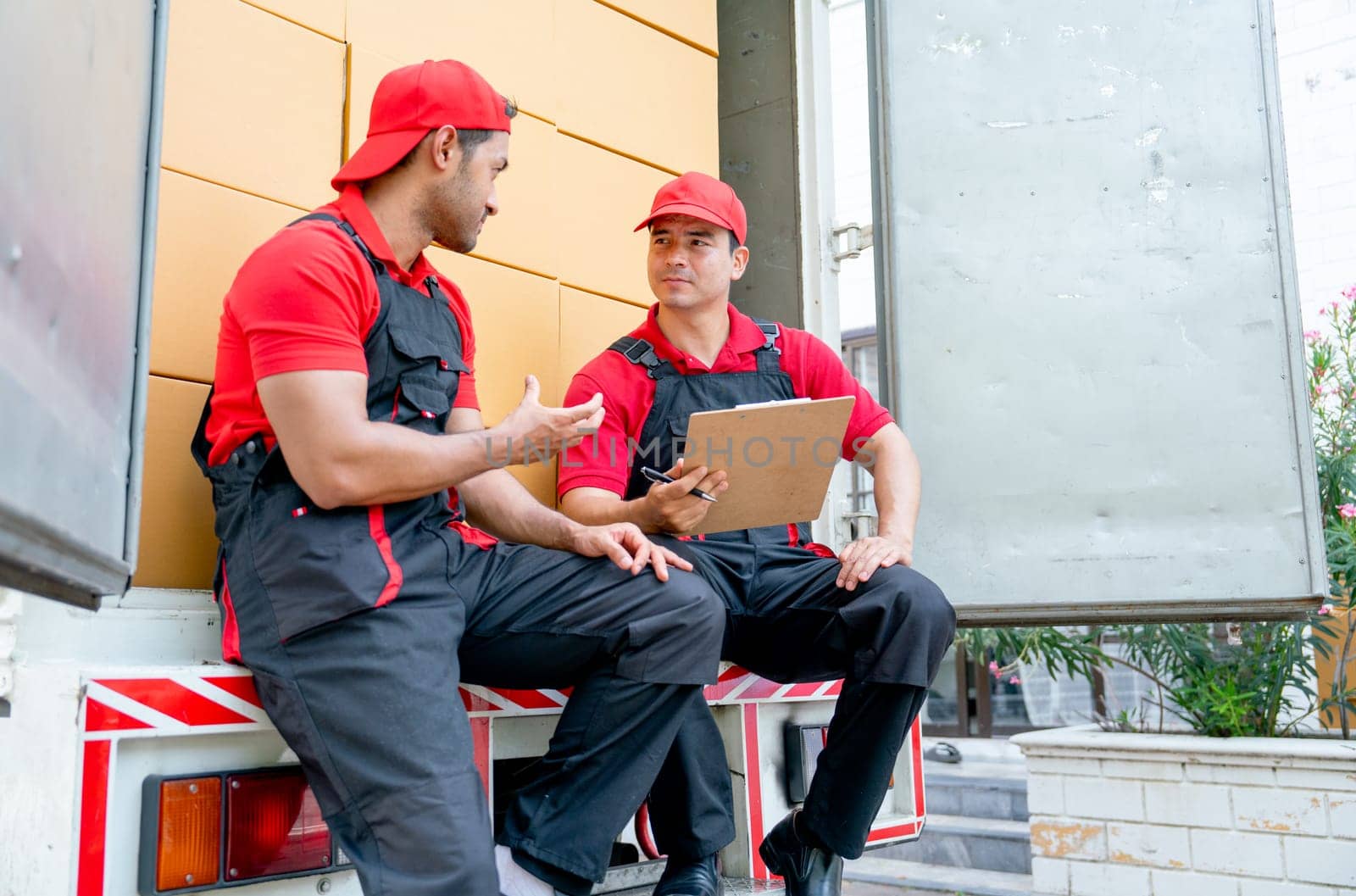 Two delivery men sit and discuss together on back part of truck during move package or boxes to customer house.