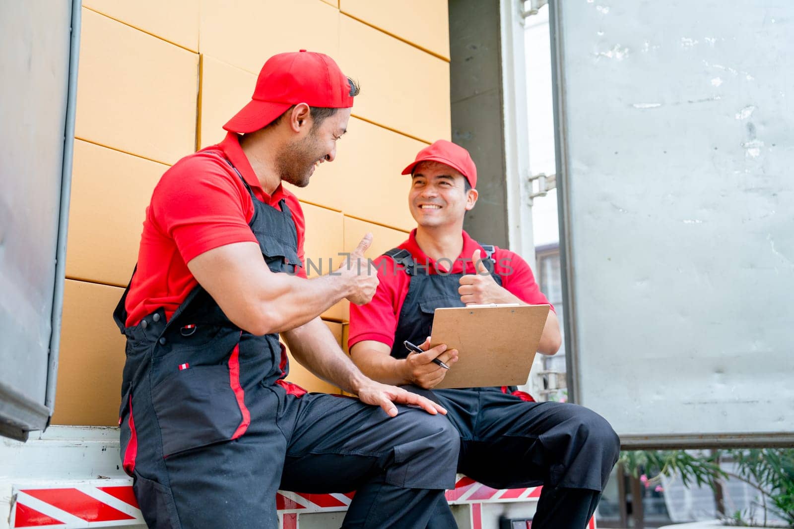Two delivery men sit and discuss together also show thumbs up on back part of truck during move package or boxes to customer house.