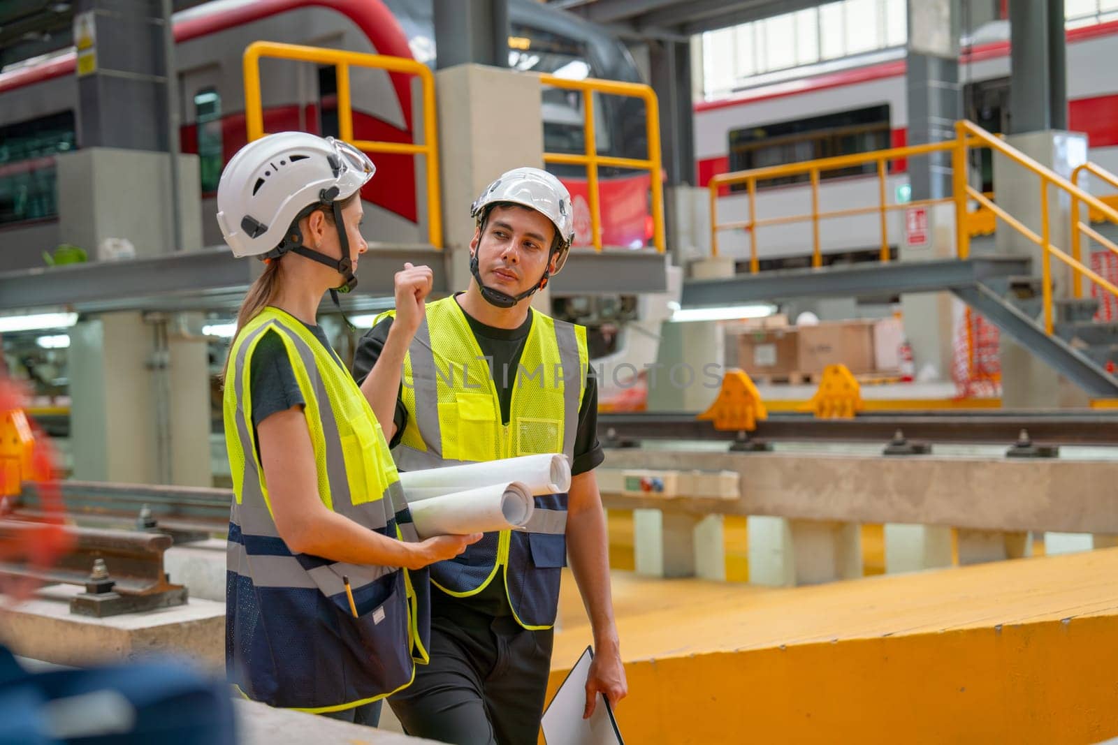 Side view of train factory workers man and woman discuss about work in workplace of maintenance center with electric train on rail is in the background.