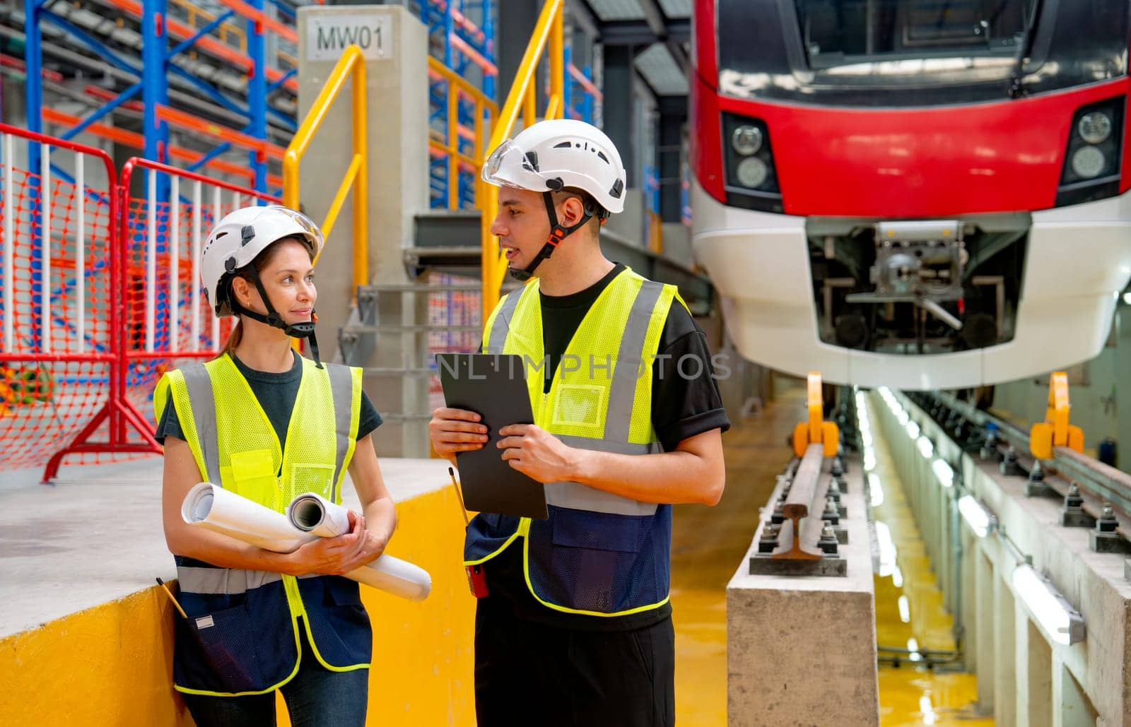 Train factory workers man and woman that one hold document pad discuss about the project in workplace with electric train is in the background.