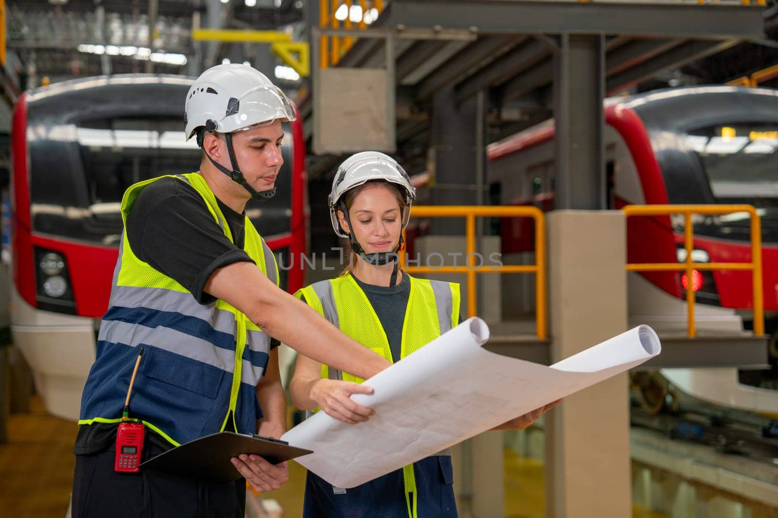 Two professional engineer or technician workers man and woman concentrate to look the information in drawing paper and they stay in front of electric train in factory workplace.